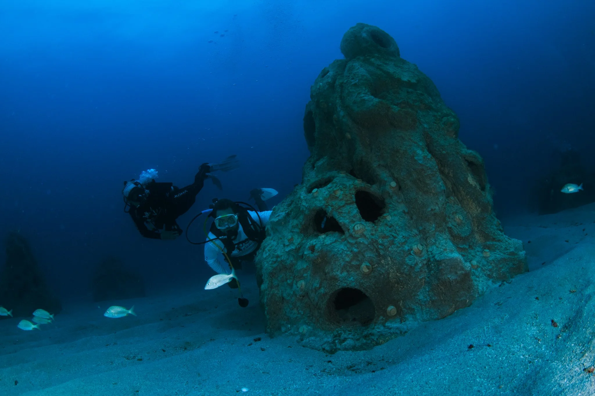 Two scuba divers swimming near a large underwater rock formation with holes, surrounded by sand and small fish.