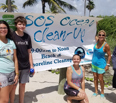 Four people standing and sitting on the beach in front of a sign that reads 'SOS Ocean Cleanup, 9:00am to noon, Beach & Shoreline Cleanup.' They are participating in an ocean cleanup event.