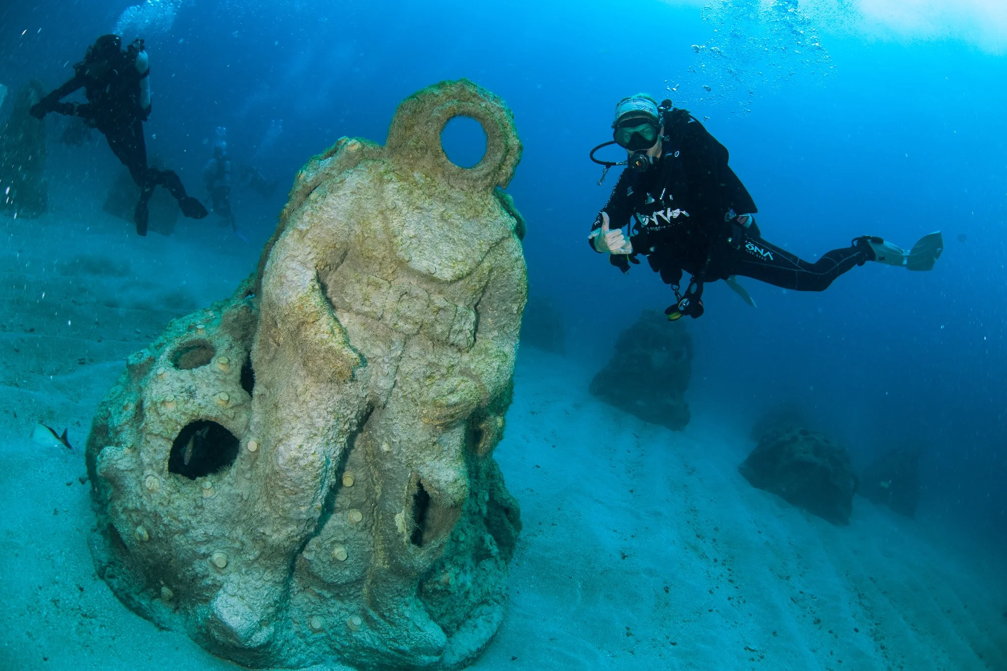 Scuba divers exploring underwater ruins with large stone sculpture of a face, surrounded by water and rocks.