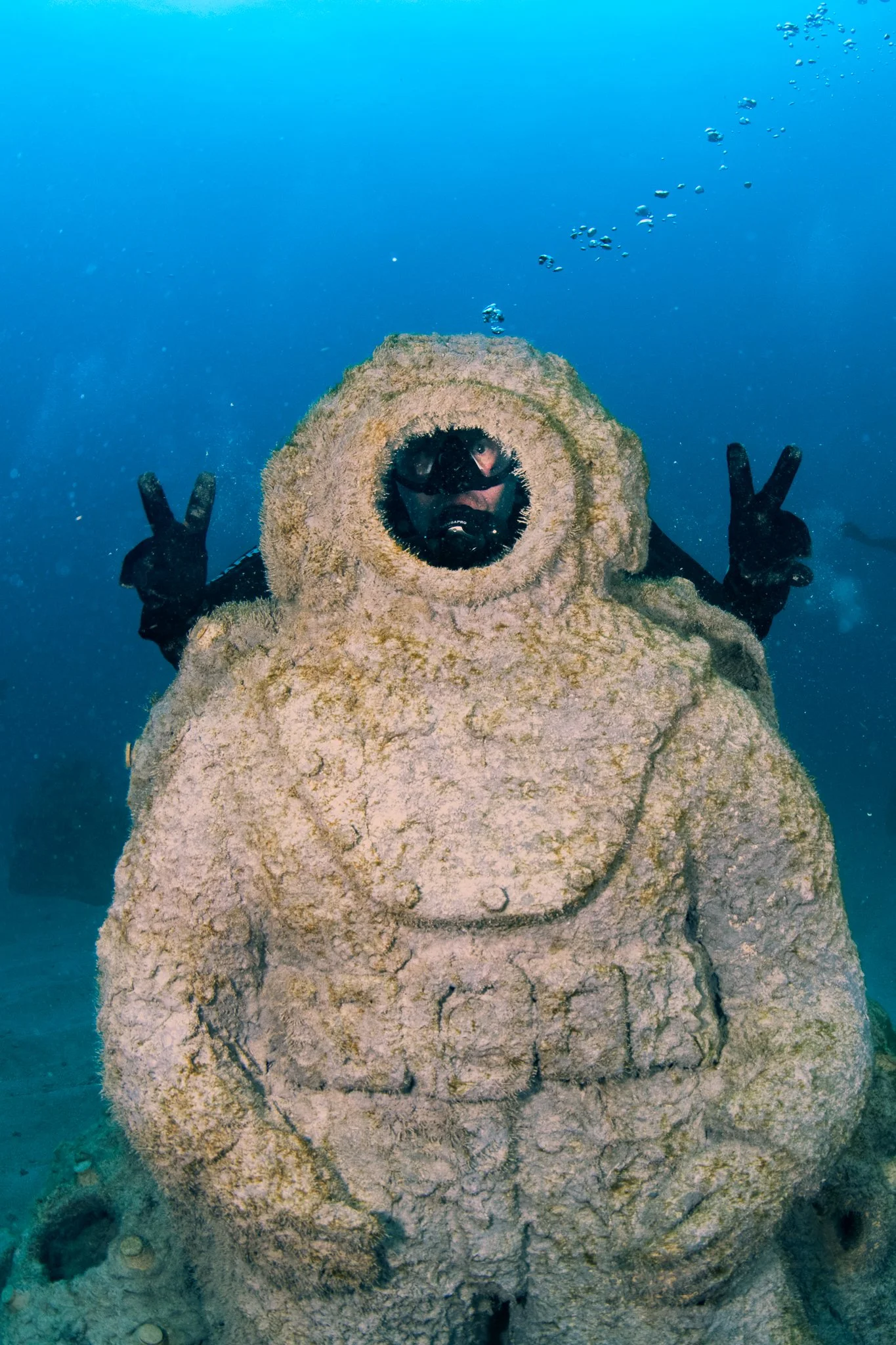 A scuba diver inside a large, stone-carved statue of a toad or frog underwater, with hands raised in a peace sign.