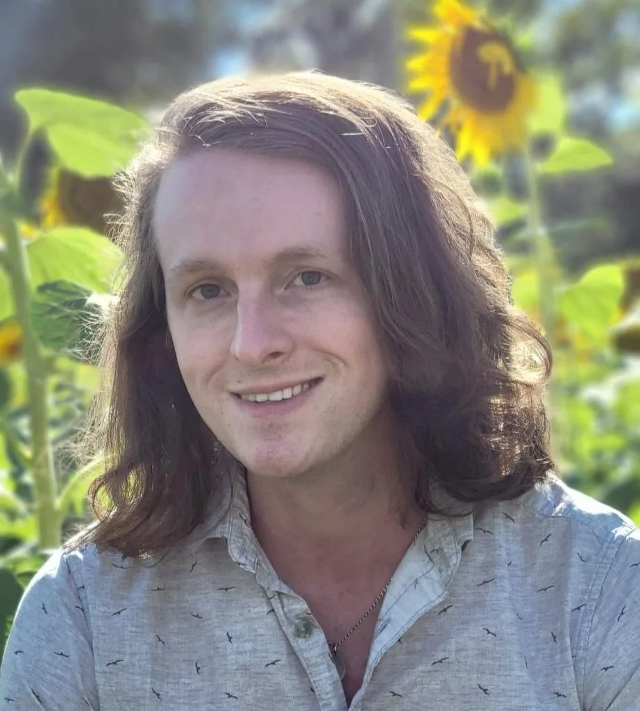 A man with long brown hair smiling outdoors, with sunflowers in the background.