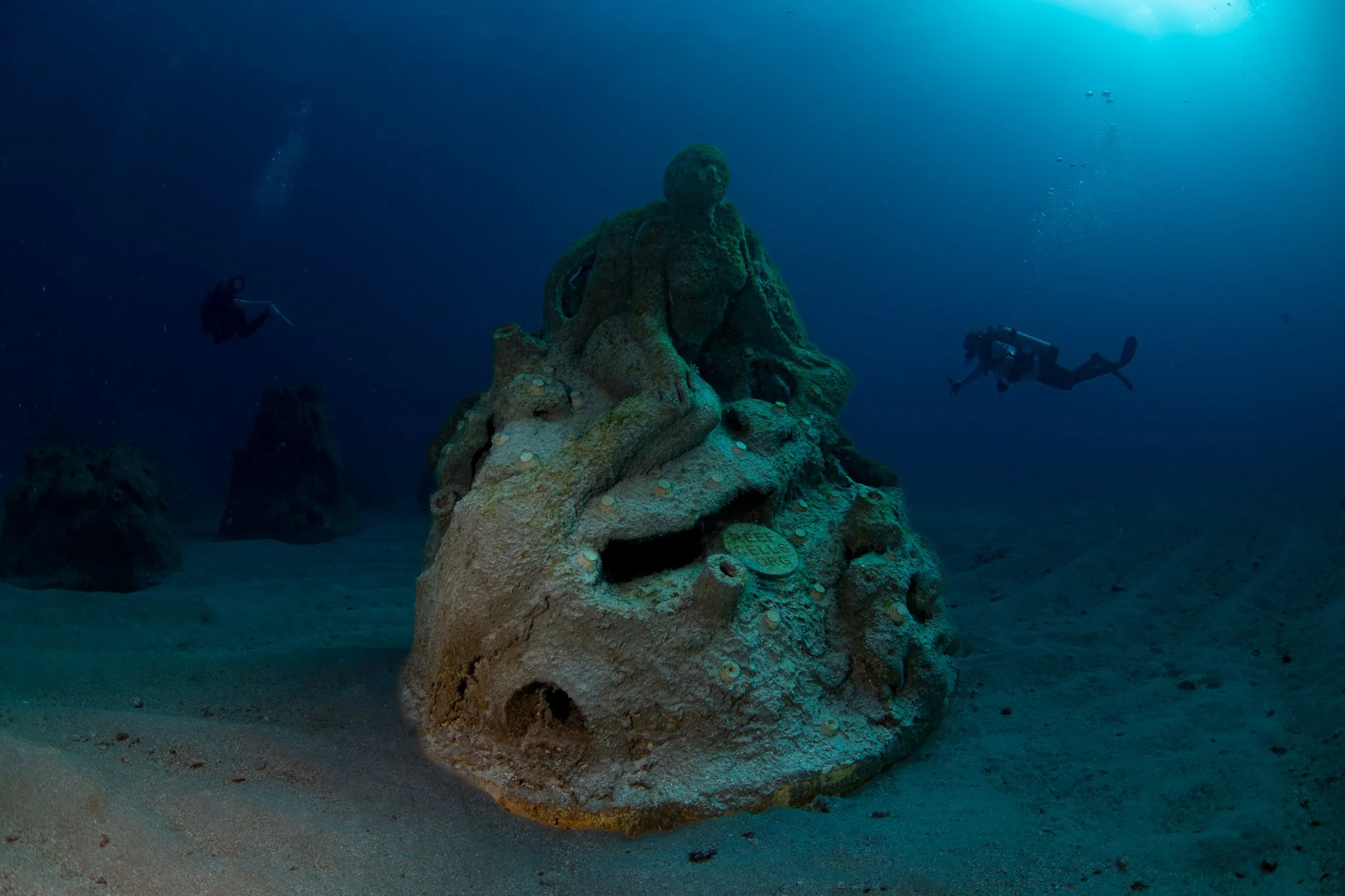 Underwater scene with scuba divers swimming around a large, weathered automatic teller machine (ATM) submerged on the ocean floor.