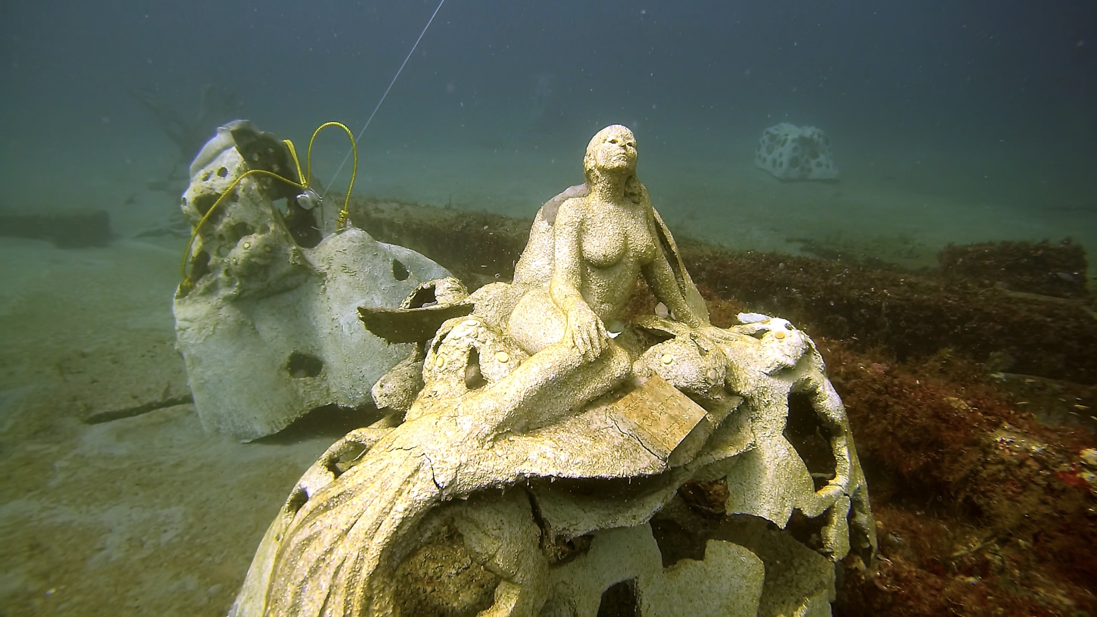 Underwater sculpture of a woman seated with her hands on her knees, surrounded by coral formations, with debris and rocks in the background.