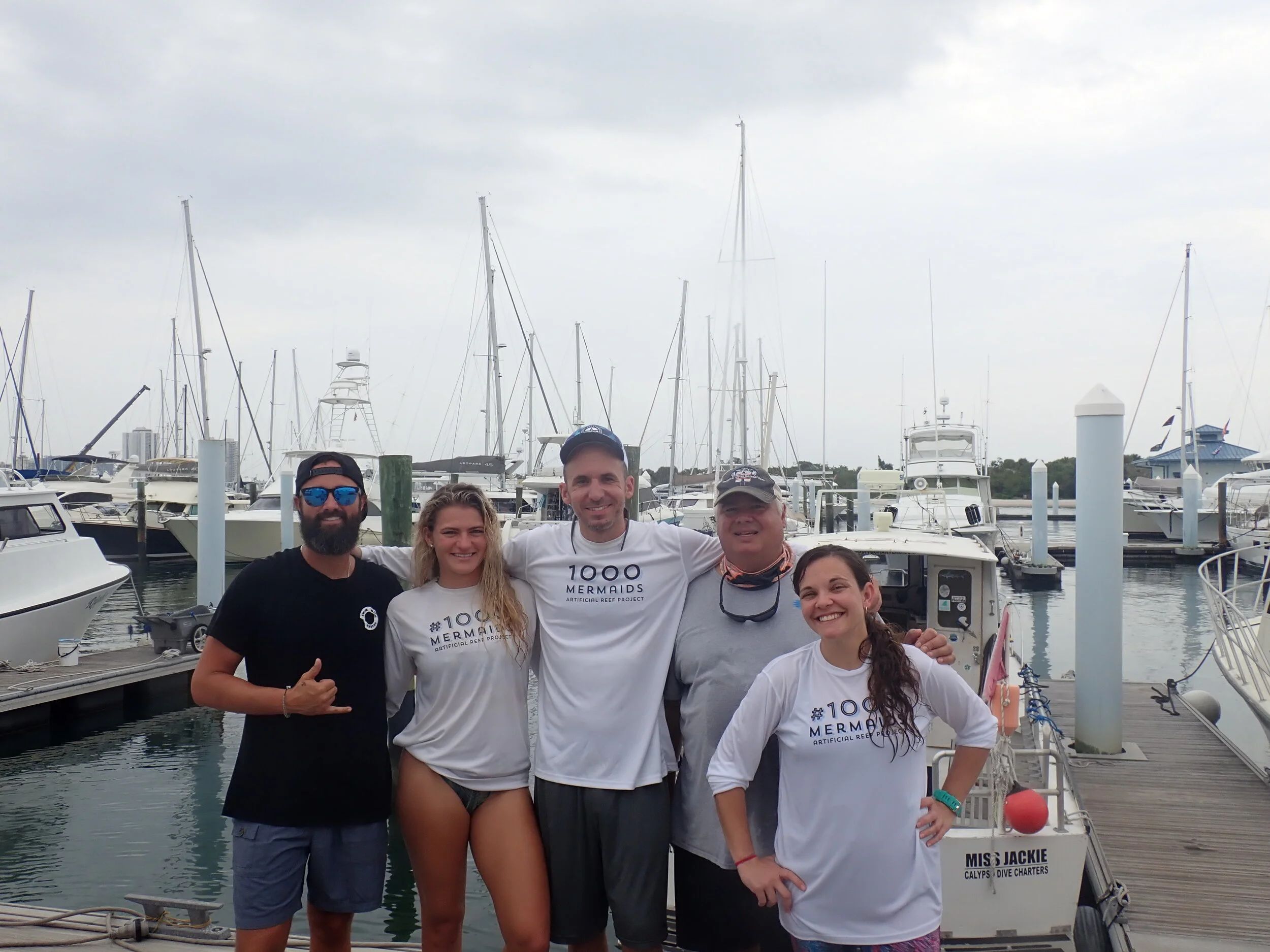 Group of five people smiling at the camera on a dock with boats and sailboats in the background.
