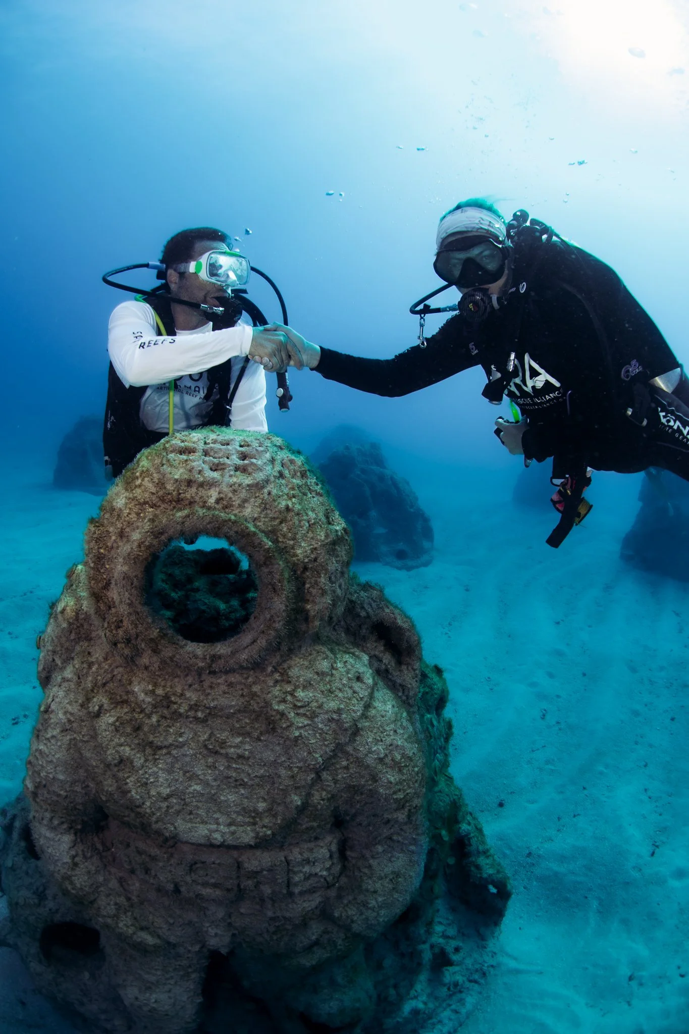 Two scuba divers underwater shaking hands near a large rusted anchor.