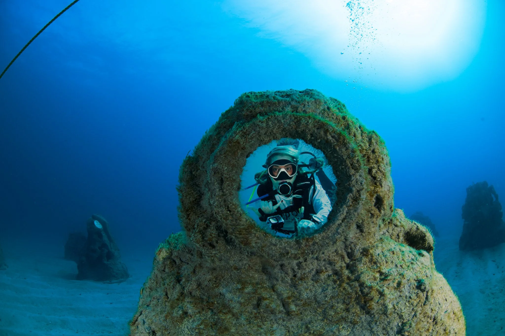 A scuba diver underwater framed through a large, circular coral-covered structure, with blue ocean background and other rock formations visible.