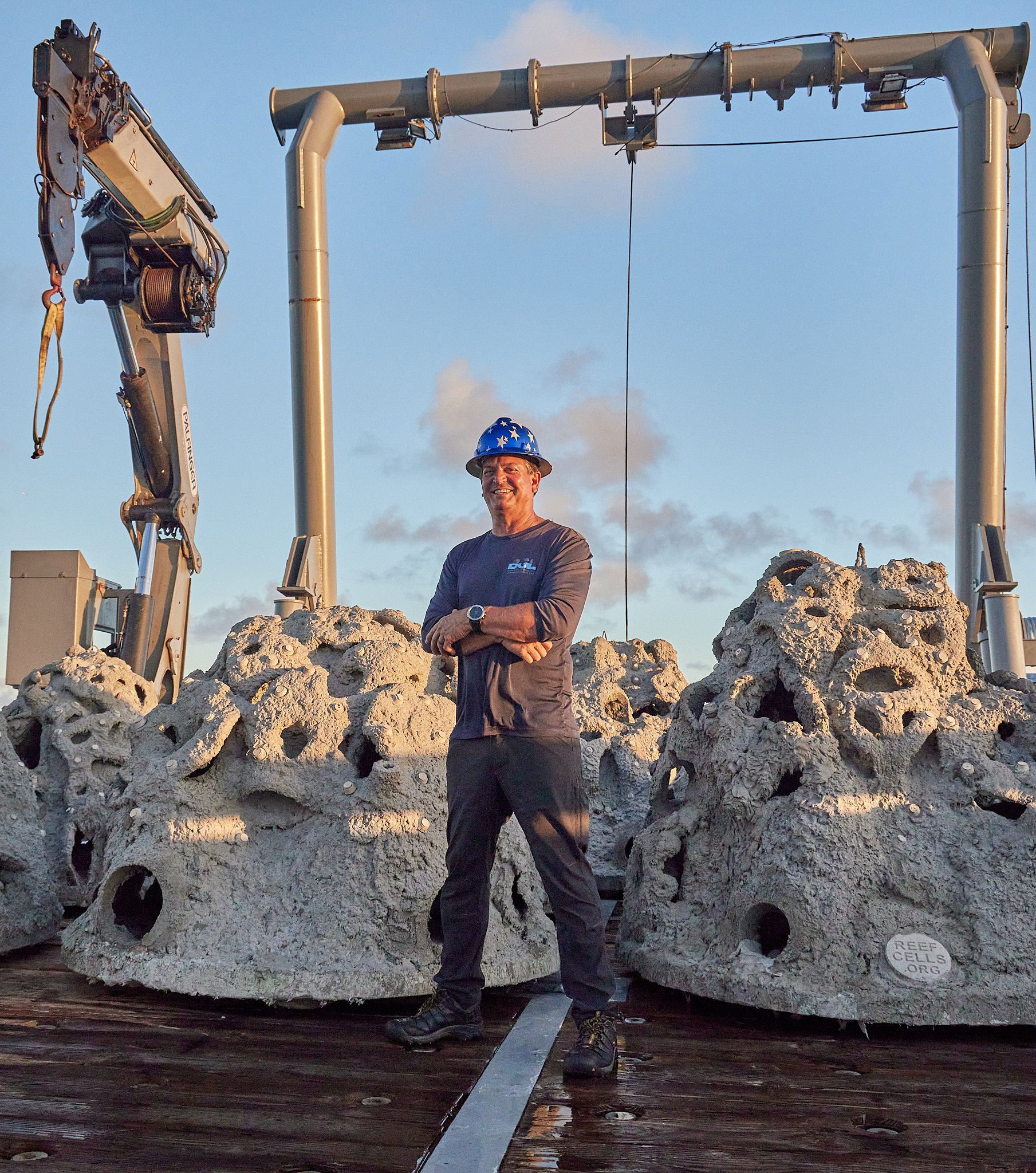 A man wearing a blue star-patterned hard hat and casual work clothes, standing on a platform surrounded by large coral rocks, with construction equipment and a metal frame structure behind him, during sunset.