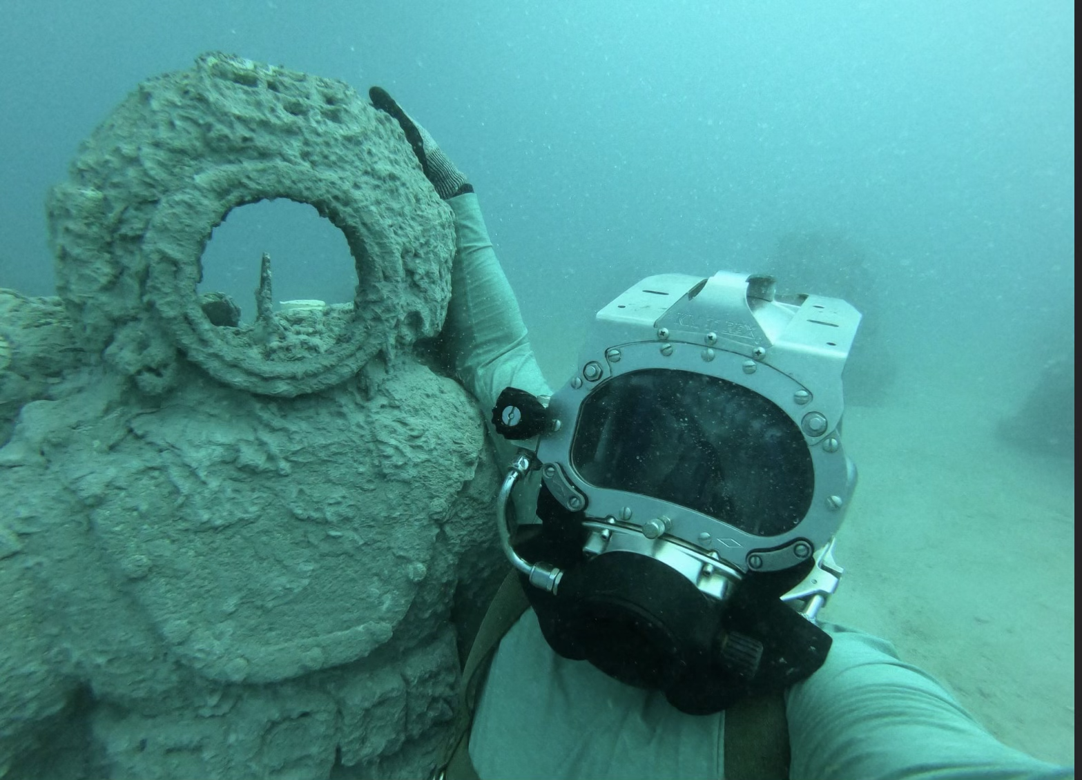 Underwater scene with a diver holding a camera, resting on a large, weathered concrete structure that appears to be part of a sunken object or wreckage.