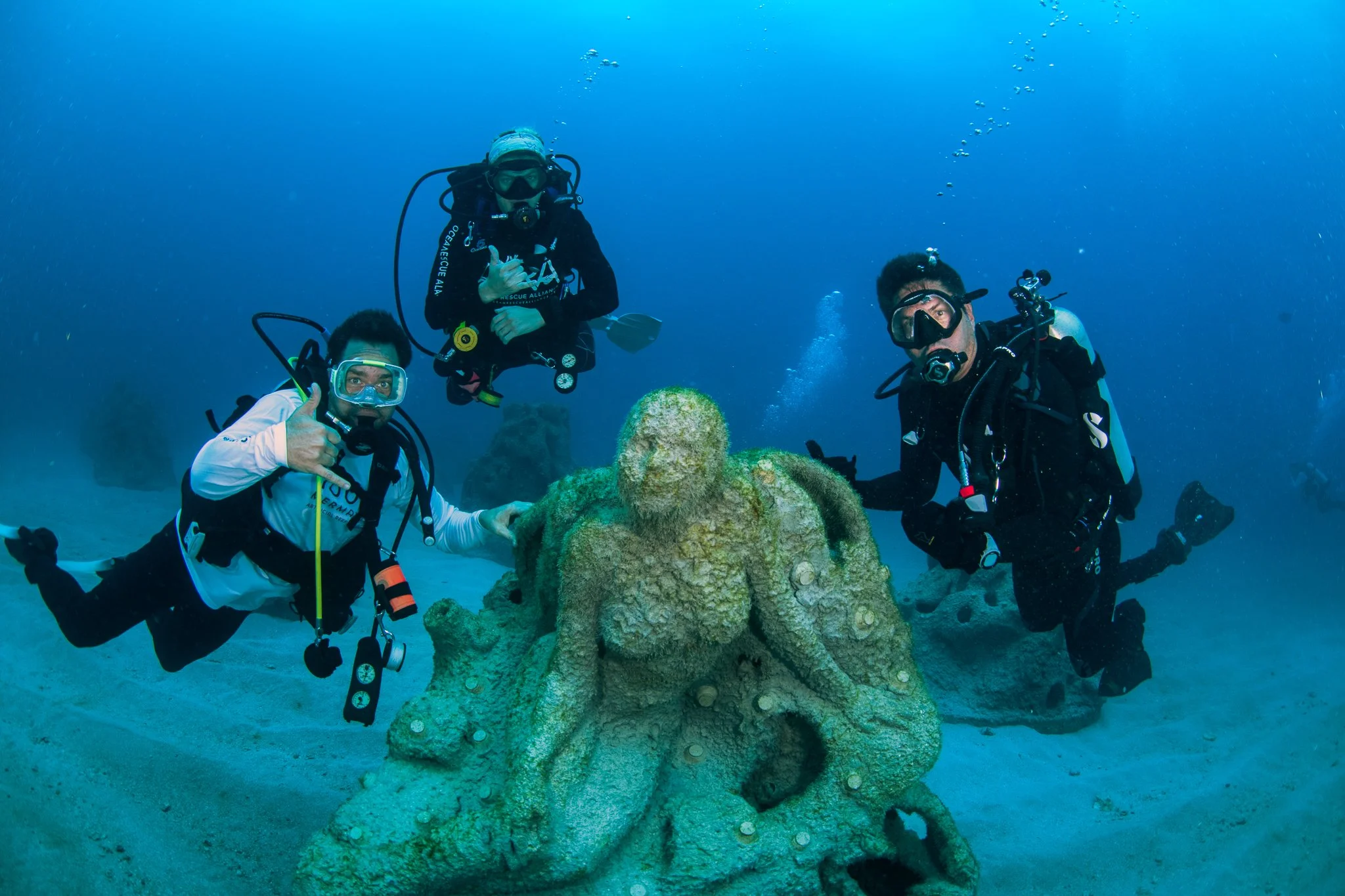 Three scuba divers underwater around a large, coral-covered octopus statue on the ocean floor.