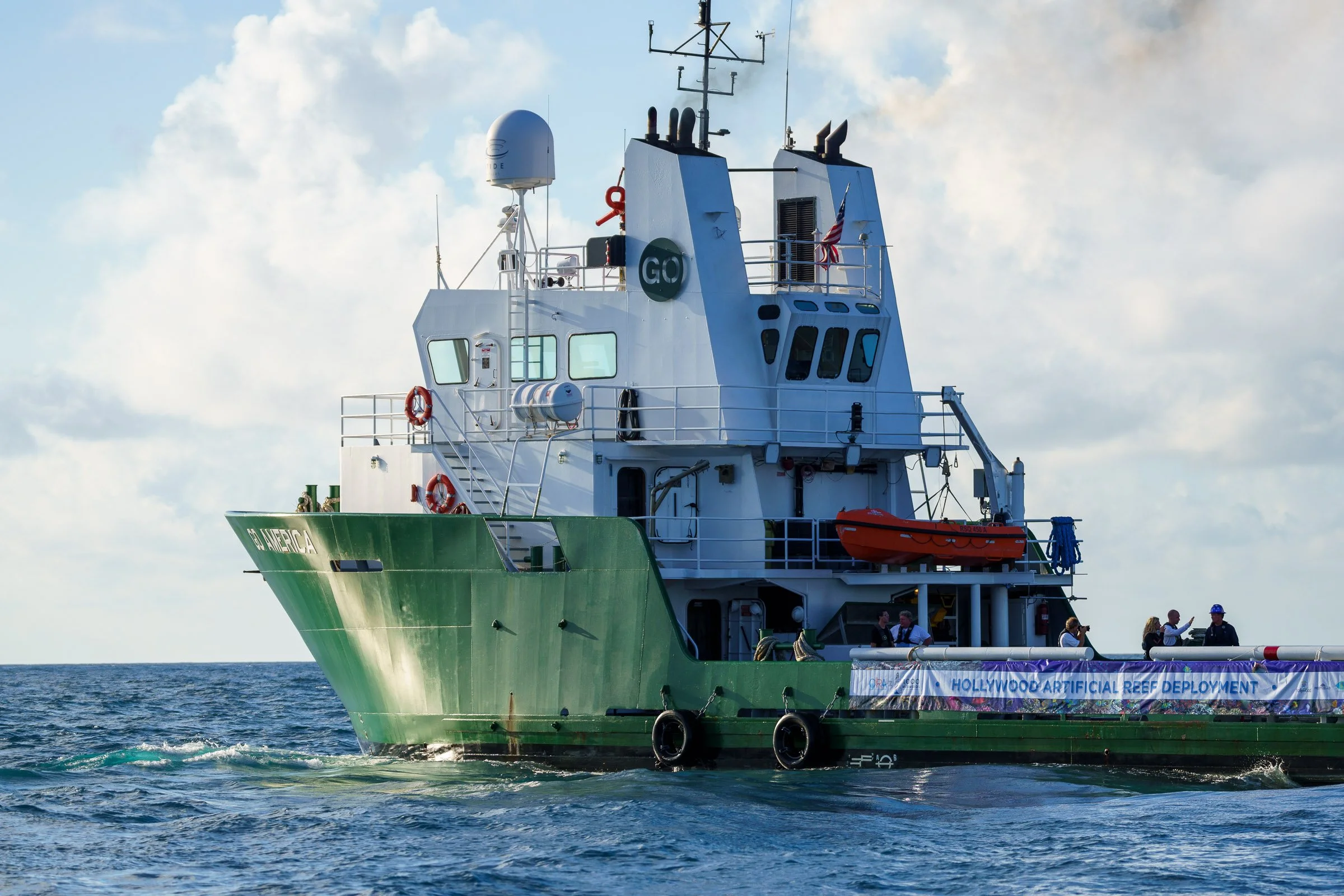 A ship with a green hull and white superstructure on the ocean, with a banner reading 'Hollywood Artificial Reef Deployment' and several people on deck.