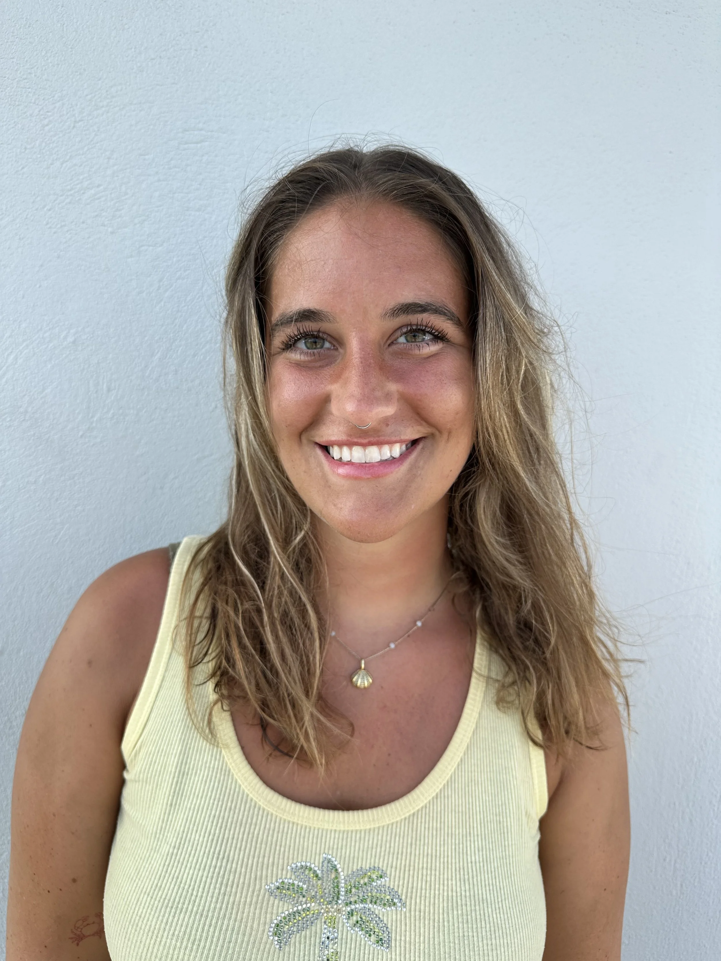A smiling woman with wavy brown hair, wearing a yellow tank top with a glittery starburst design, standing against a plain white wall.