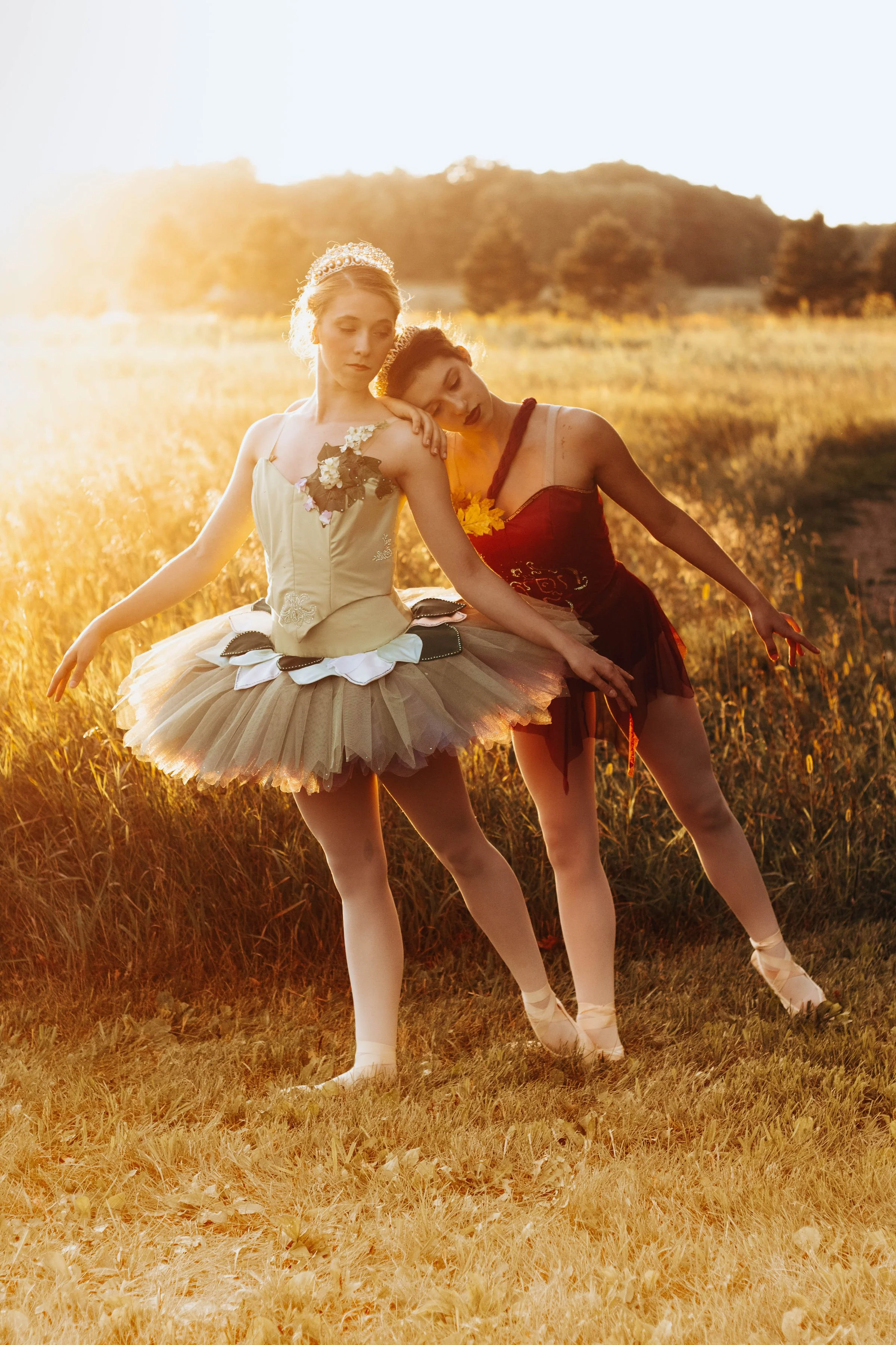 Two ballet dancers in tutus perform outdoors at sunset, with trees and a hill in the background.