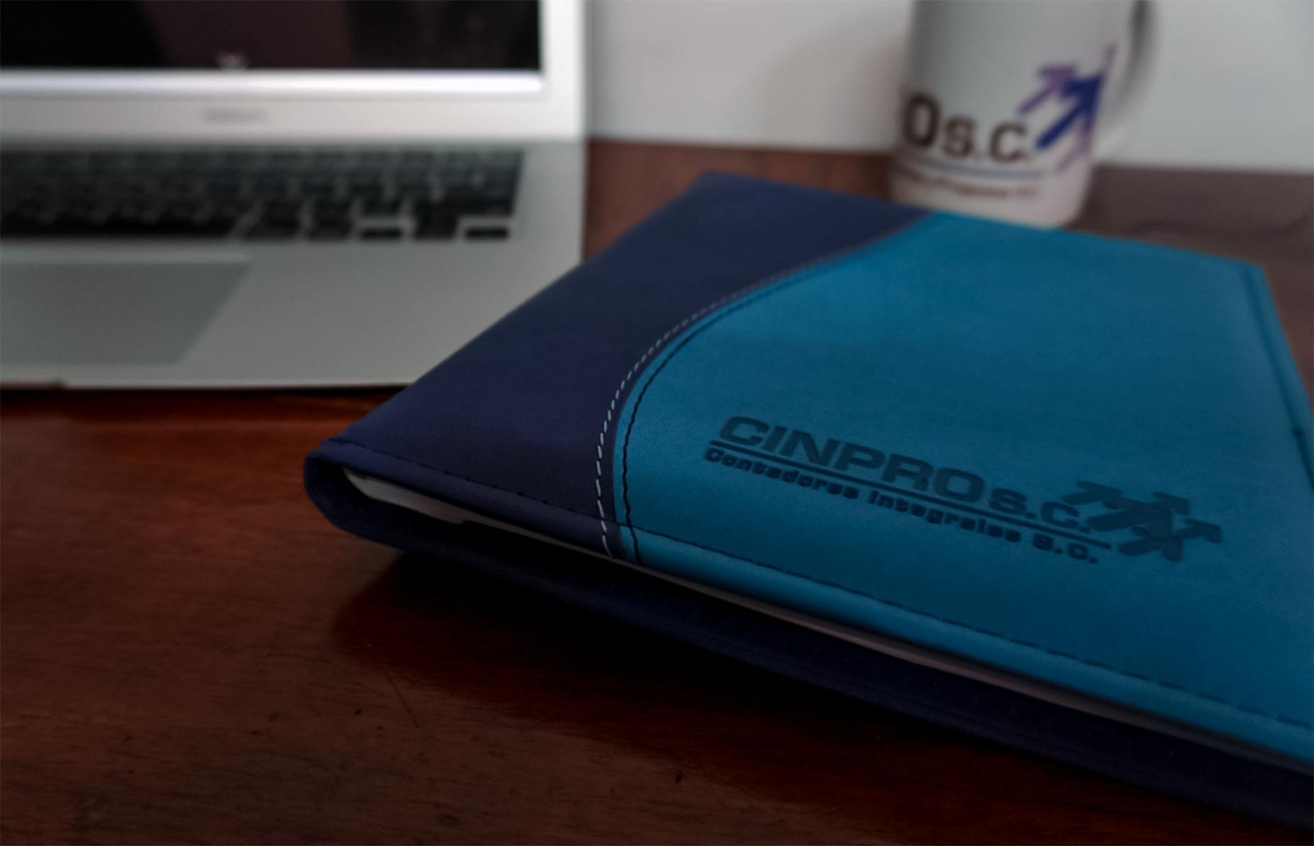 Cuaderno azul con logo y texto del colegio CNCPro en primer plano, sobre una mesa de madera. En el fondo, una laptop y una taza con logotipo y flechas. La imagen está ligeramente desenfocada.