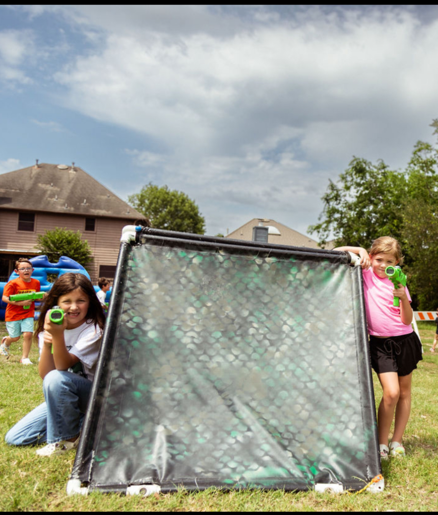 Children playing with water guns during a water fight in a backyard, with a black and transparent water barrier at the center, a house with a dome-shaped roof in the background, and cloudy sky above.