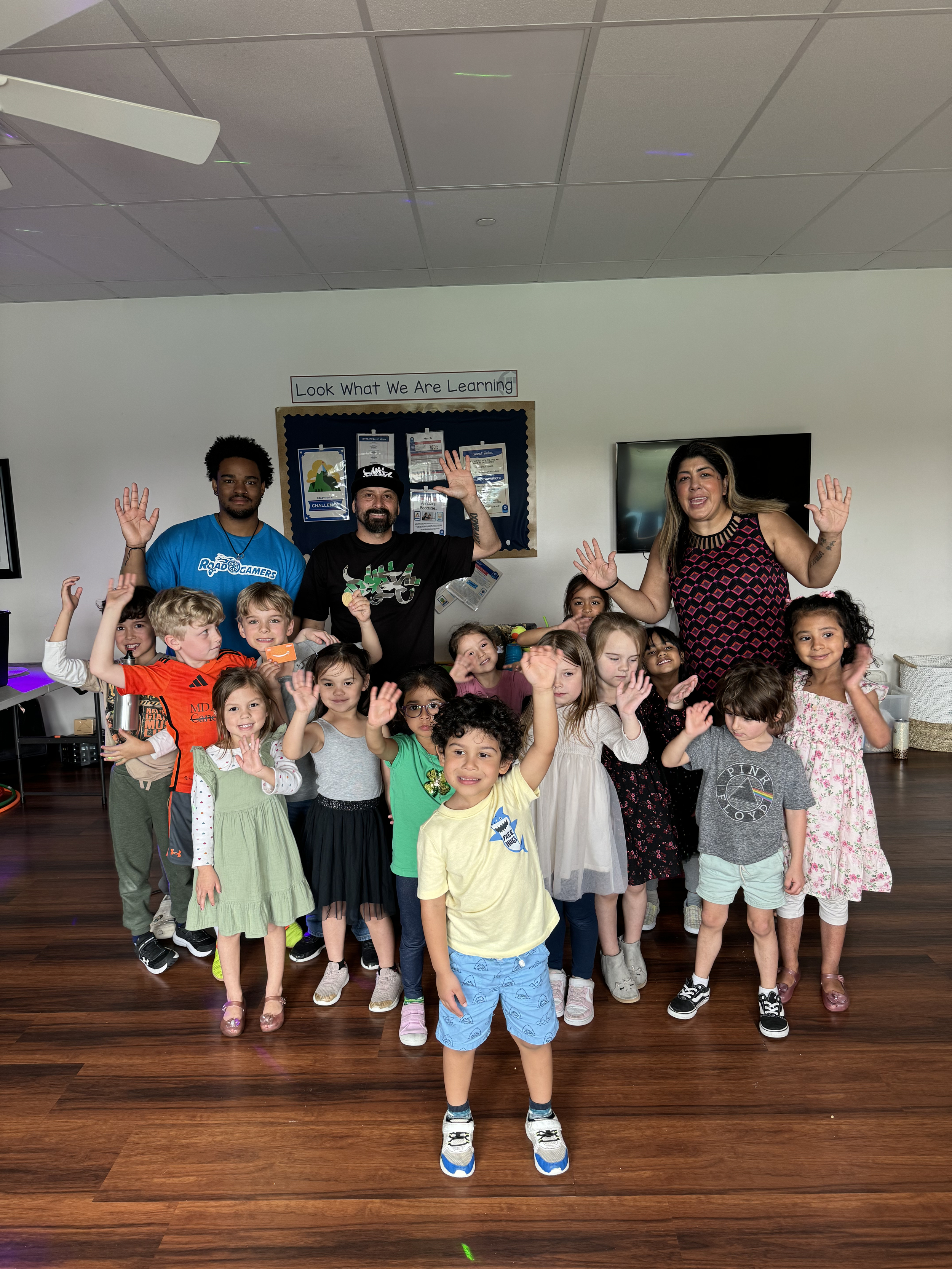 Group of children and adults smiling and raising their hands in a classroom, with a sign that reads 'Look What We Are Learning' on the wall behind them.
