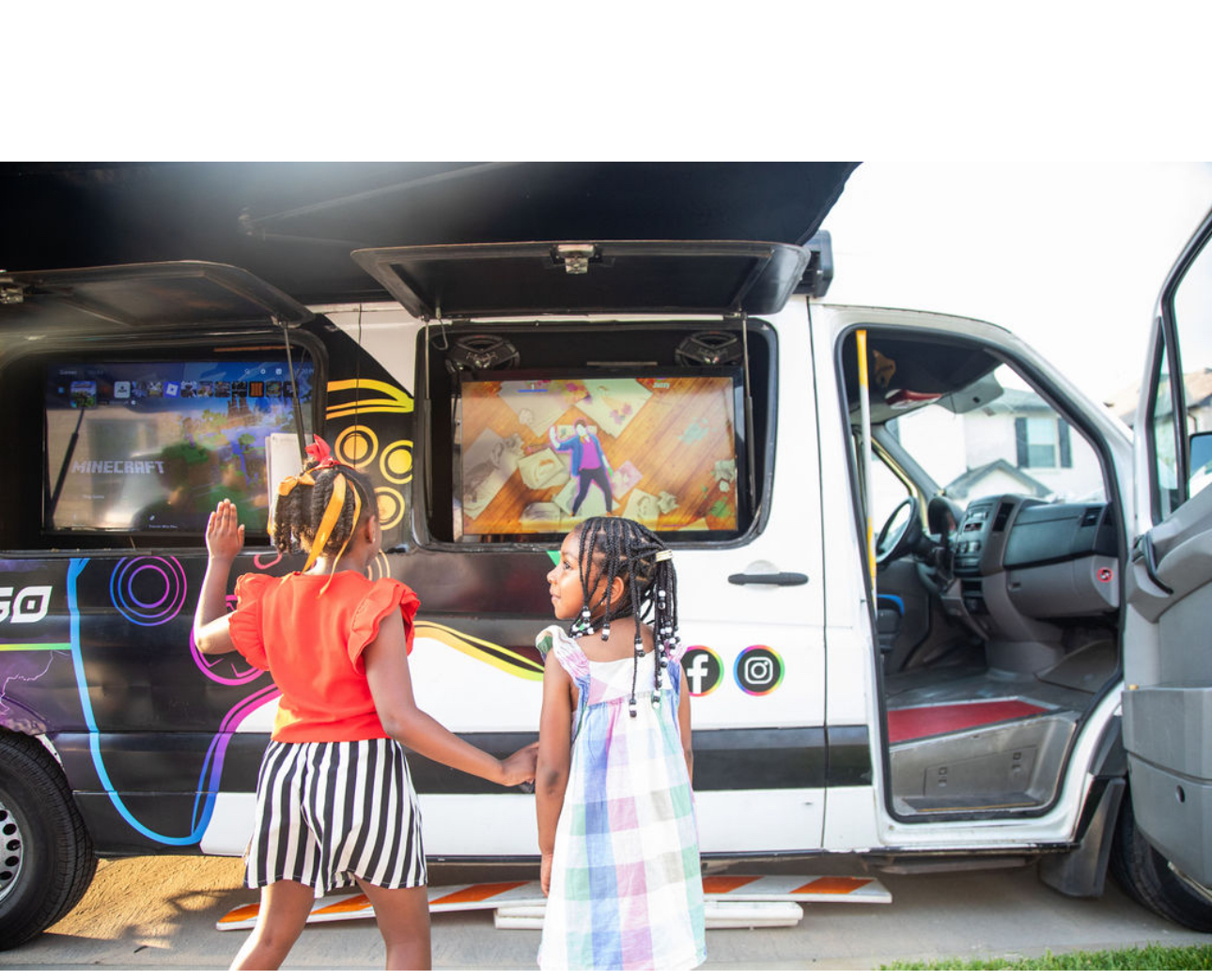 Two young girls playing video games on large screens inside a mobile gaming van, with one girl raising her hand and holding the other's hand, outside on a suburban street.