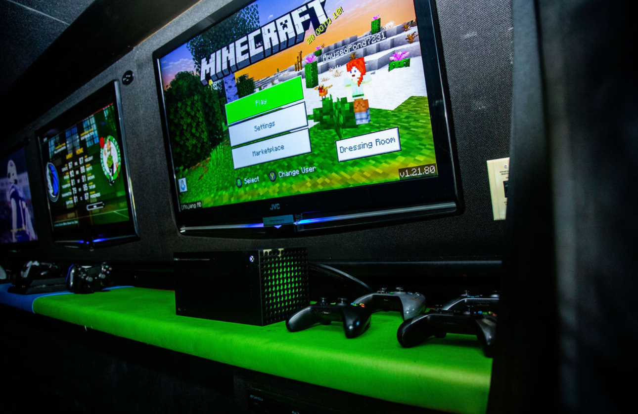 Gaming arcade with multiple screens, including a Minecraft game menu on the main screen, controllers, and gaming consoles on a fluorescent green surface.