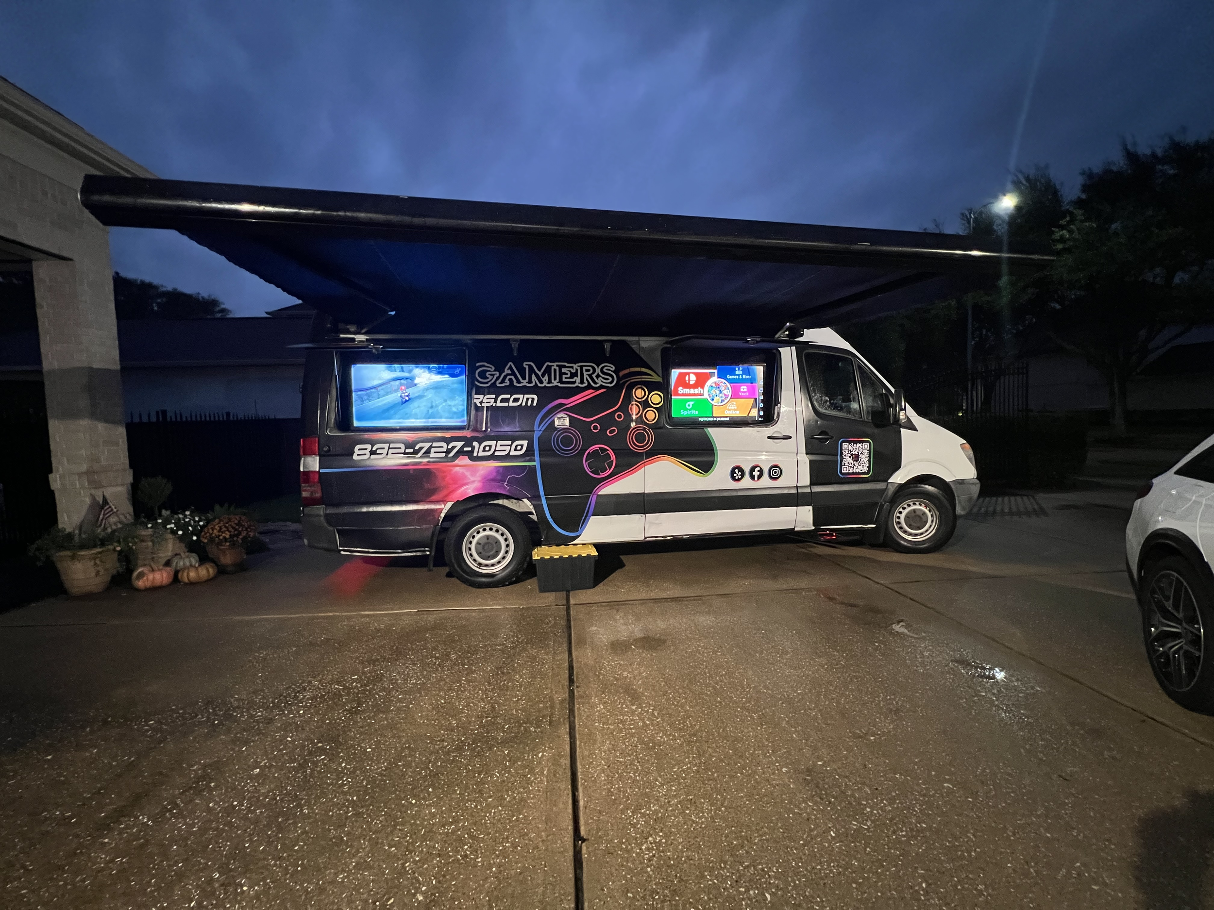 A mobile video gaming setup on a van with two screens, set up outdoors in a parking lot at dusk, with pumpkins and plants near the van.