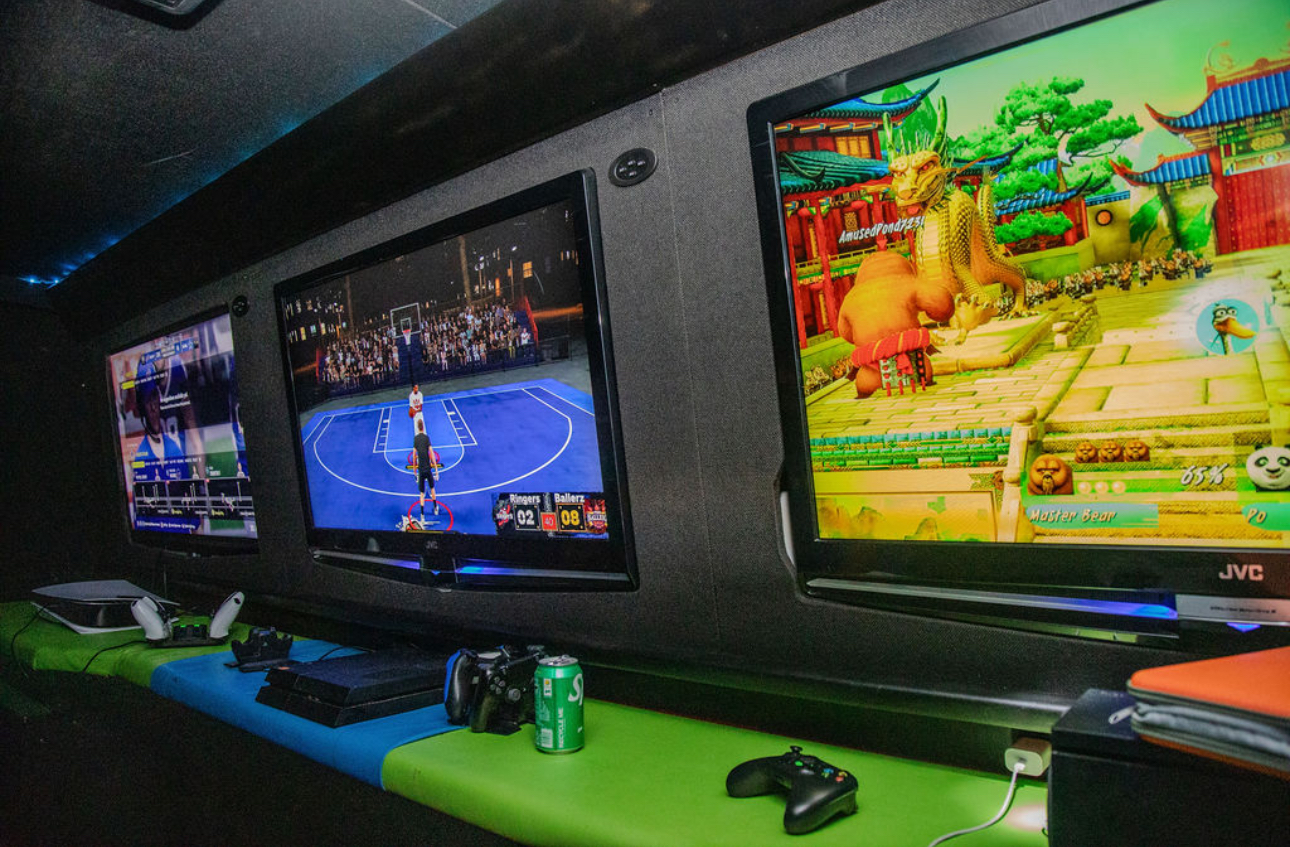 Three video game screens on a wall, with gaming controllers and a soda can on a table below.