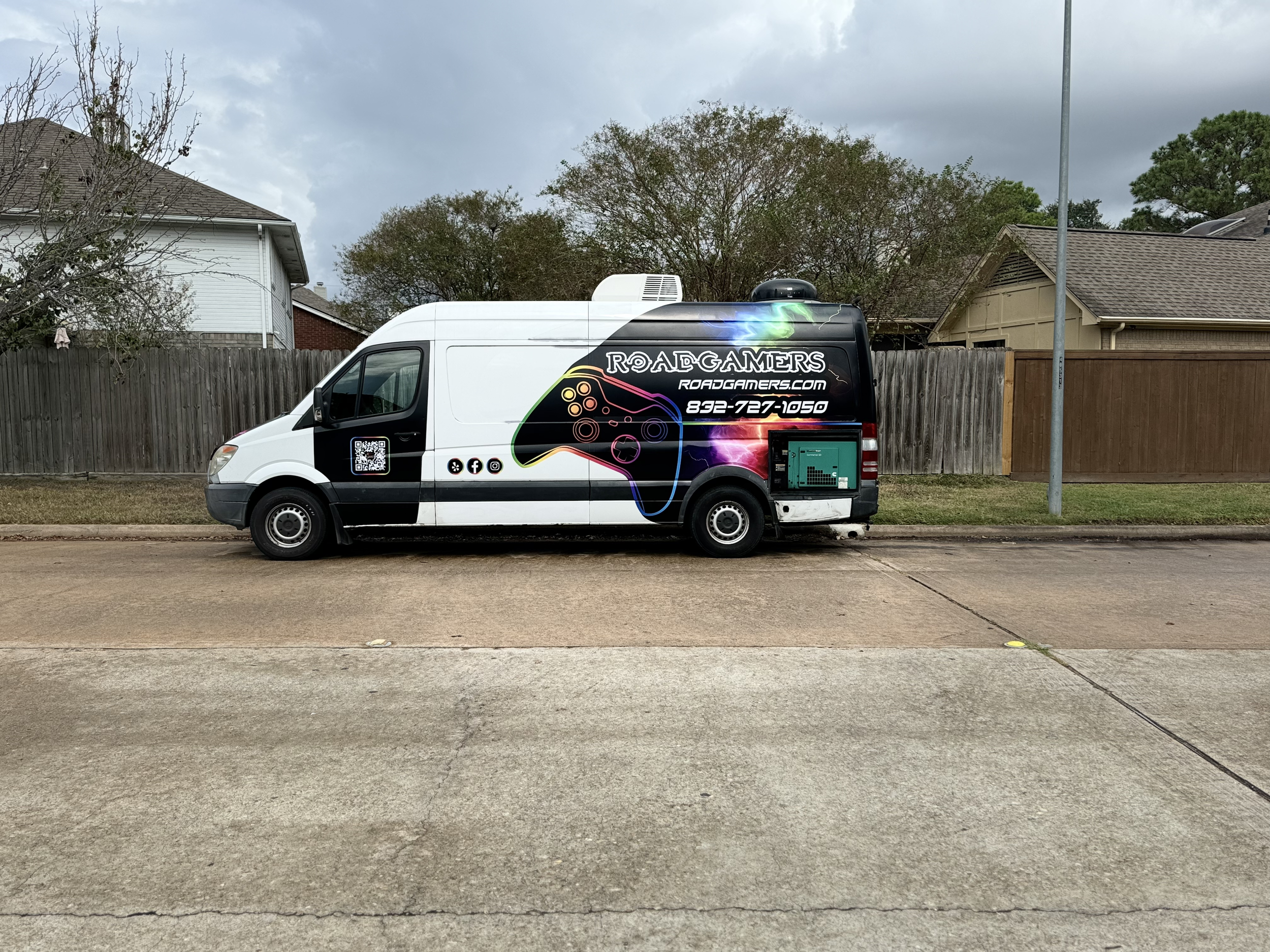 A delivery van parked on a suburban street with a black and white exterior, featuring colorful graphic of a game controller, the text 'Roadgamers,' website 'roadgamers.com,' a phone number, and social media icons.