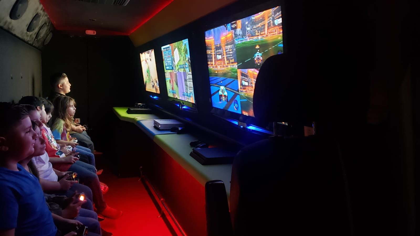 Children sitting in a dark arcade room, playing video games on large screens using gaming controllers.