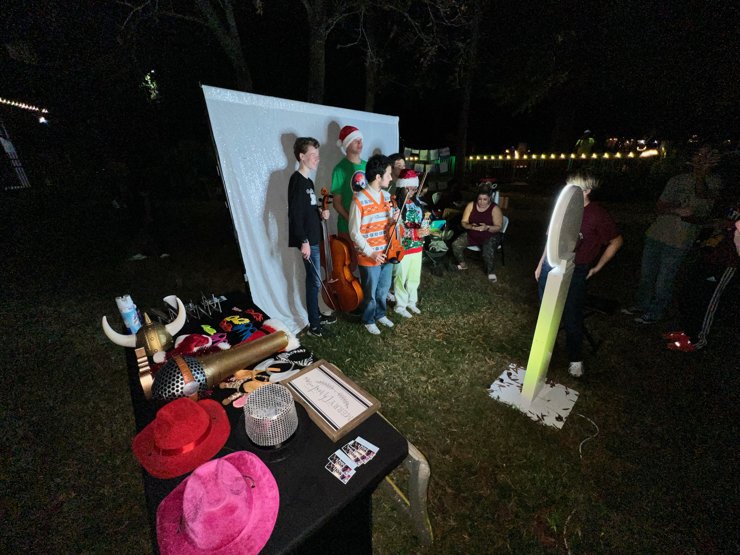 Group of people performing on a small outdoor stage at night, with props and decorative items in front, including hats and photo booth accessories, some people dressed in Christmas themed attire, and an illuminated standing mirror.