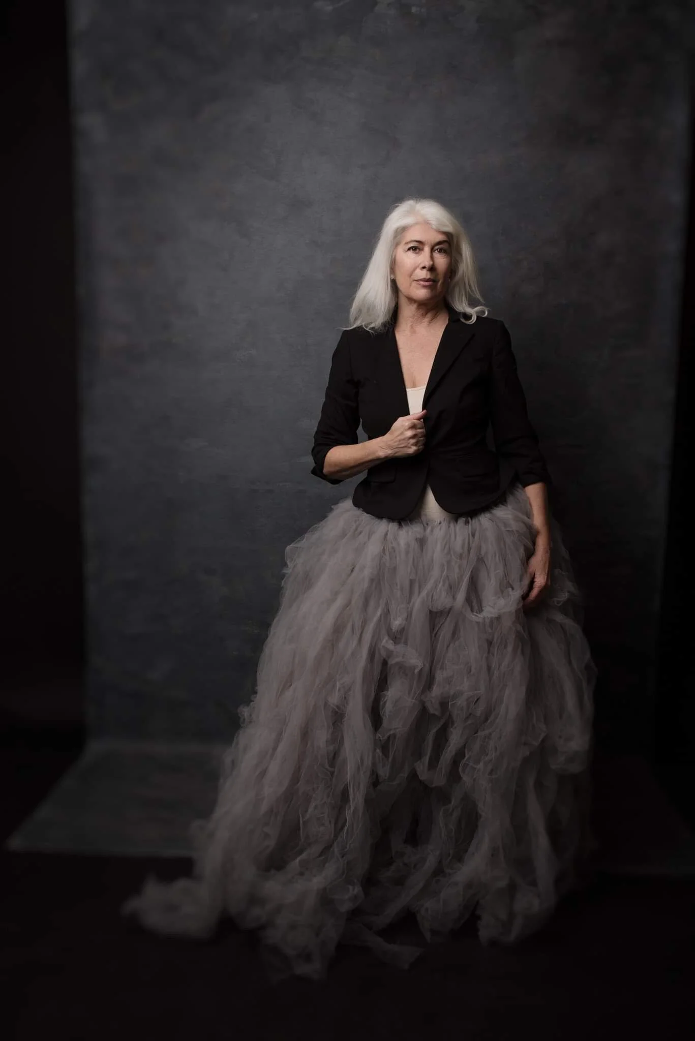 An older woman with silver hair standing against a dark textured background, wearing a black blazer over a white top and a voluminous, layered beige tulle skirt.