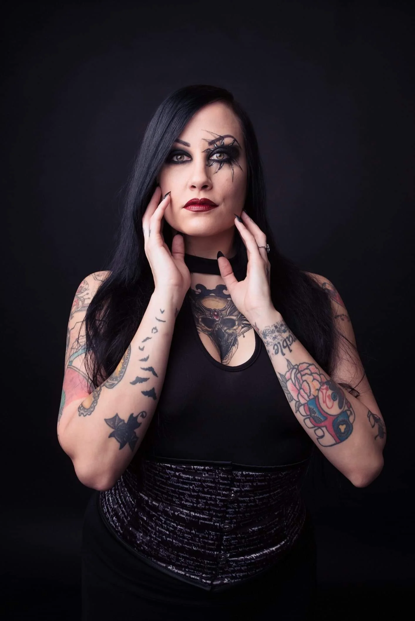 A woman with long black hair and tattoos on her arms, wearing dark makeup, a black top, and a dark background.