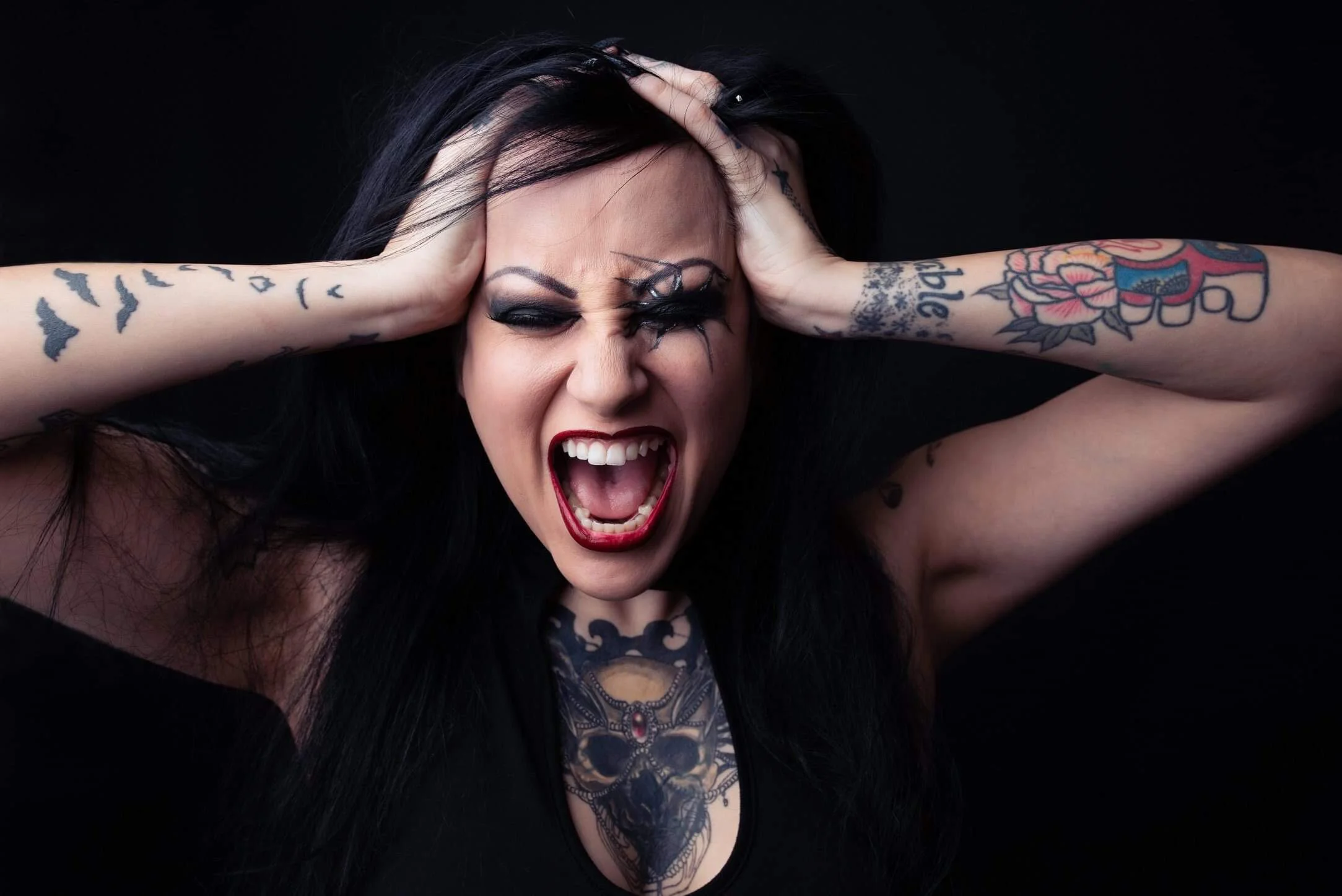 A woman with tattoos on her arms and chest, dark hair, red lipstick, singing passionately with hands on her head against a black background.