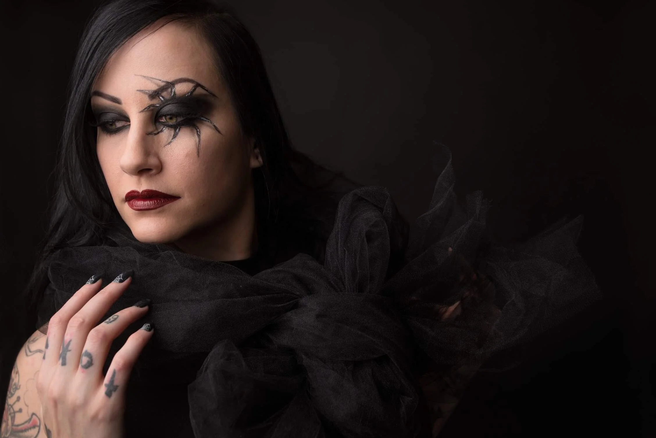 A woman with black hair and dark makeup, wearing black tulle clothing and with spider and web face paint around her eye, posing against a black background.