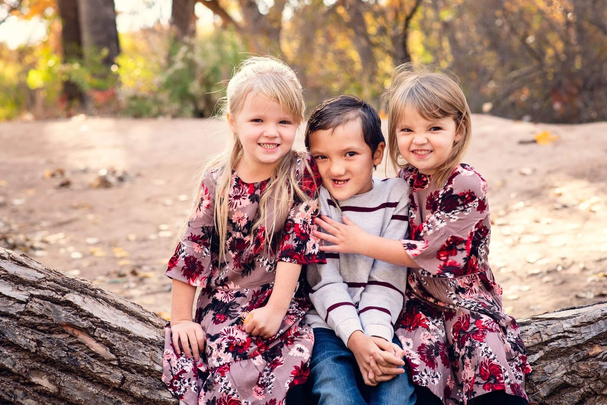 Three children, two girls and one boy, sitting on a large fallen tree in a park with autumn trees in the background. The girls are wearing floral dresses, and the boy is wearing a striped sweater. They are smiling and appear to be happy.