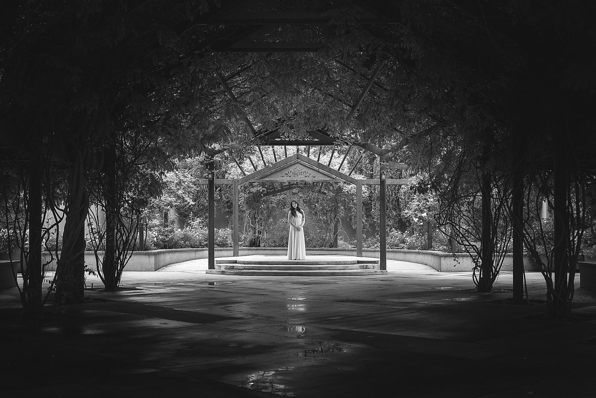 A woman standing on a circular platform in a park, under a small wooden gazebo, surrounded by trees, with a canopy overhead, in black and white.