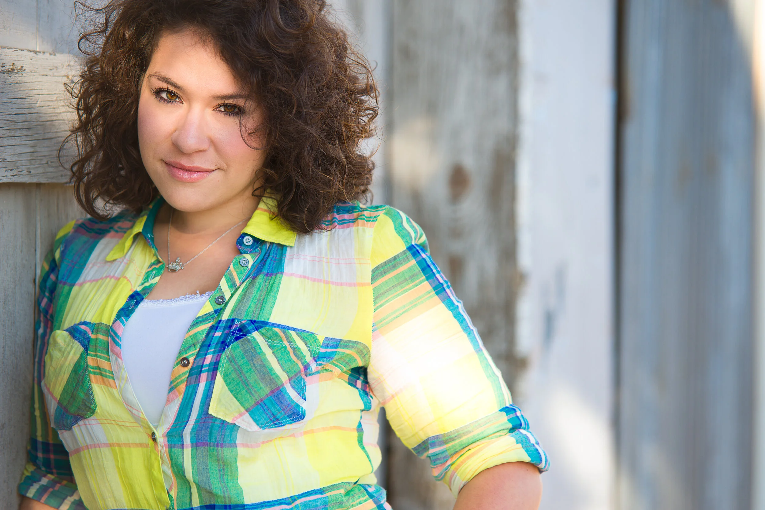 A young woman with curly brown hair leaning against a wooden fence, wearing a colorful plaid shirt with yellow, green, blue, and pink shades, and a white top underneath.