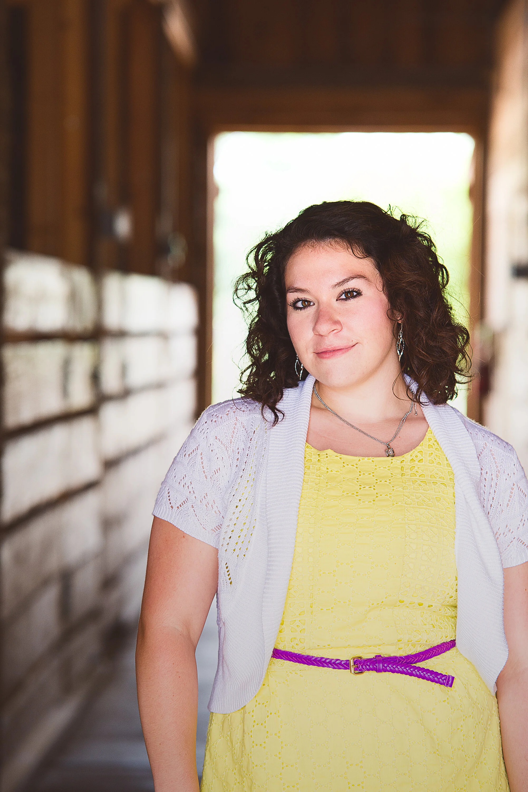A young woman with curly brown hair and makeup, wearing a yellow dress with a purple belt, silver jewelry, and a white knit shrug, standing in a rustic wooden barn corridor with daylight coming through the open door behind her.