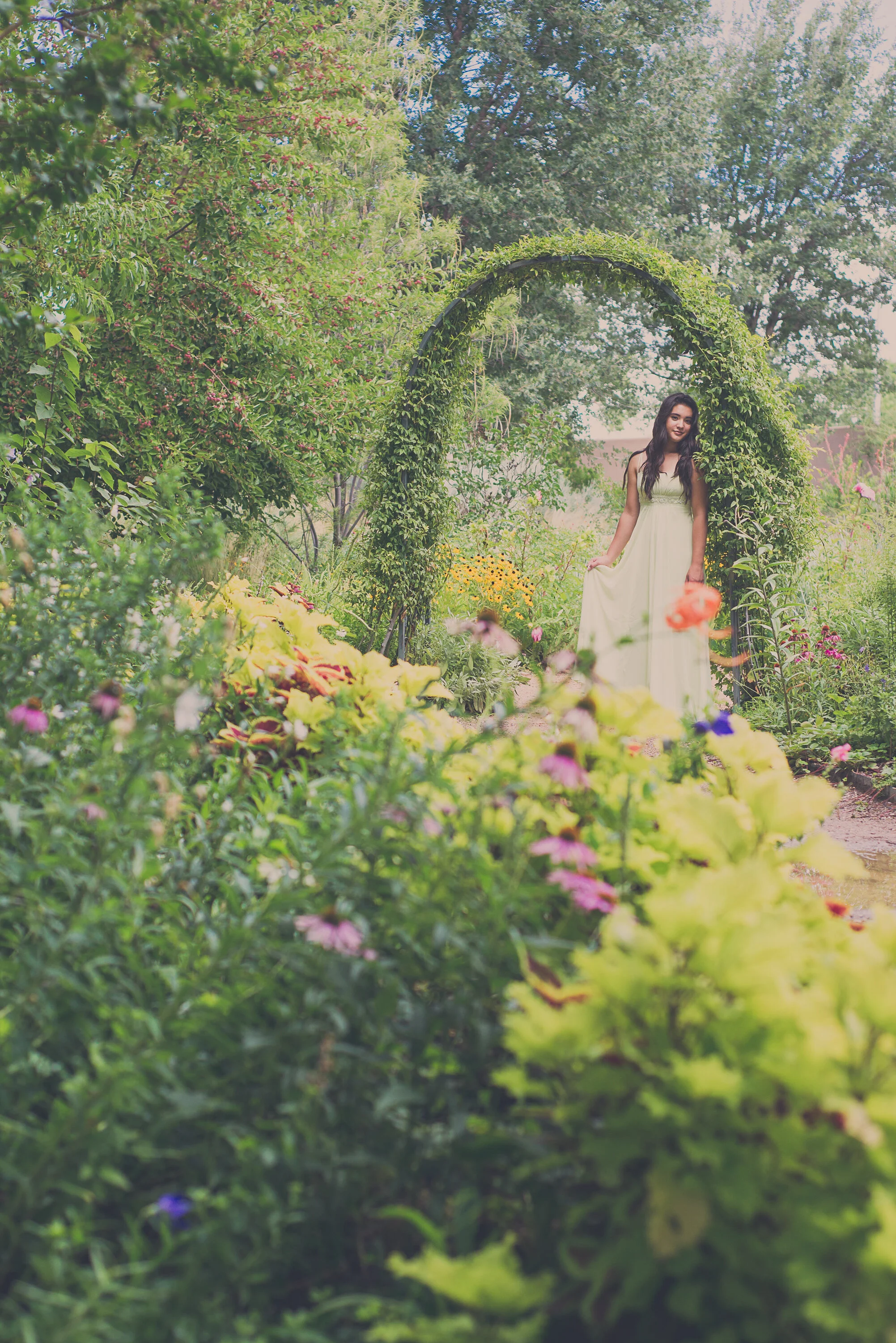 Young woman in a flowing white dress standing under a green garden arch surrounded by colorful flowers and lush trees.