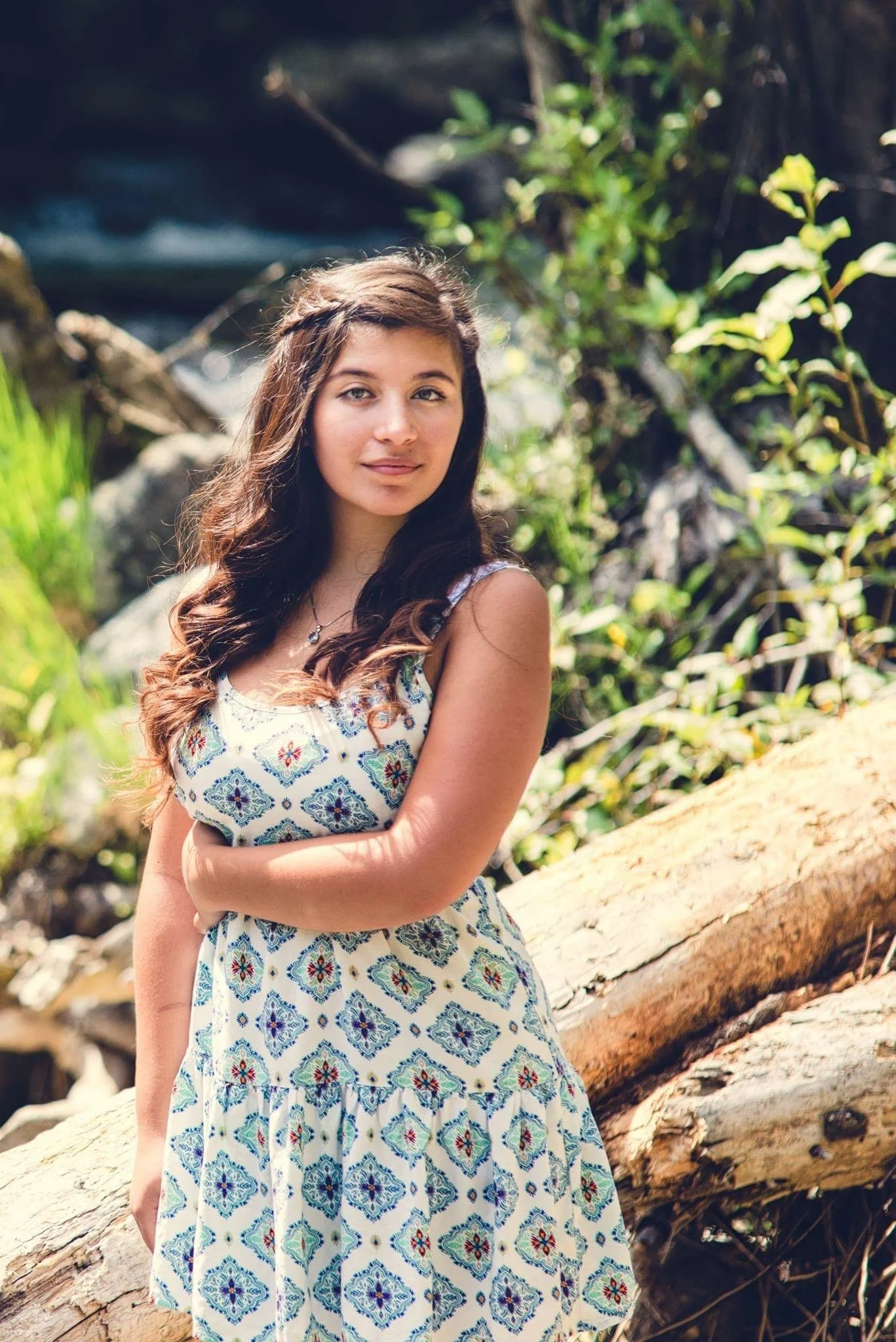 A young woman with long, wavy brown hair stands outdoors near a fallen log and green foliage, looking into the camera.