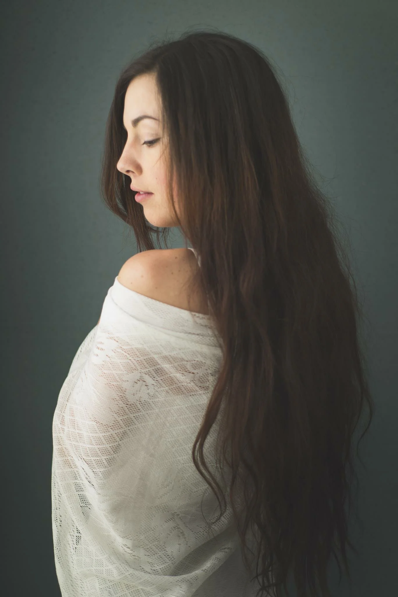A woman with long, wavy dark hair is looking to the side with her eyes closed, wearing an off-shoulder white lace top against a gray background.