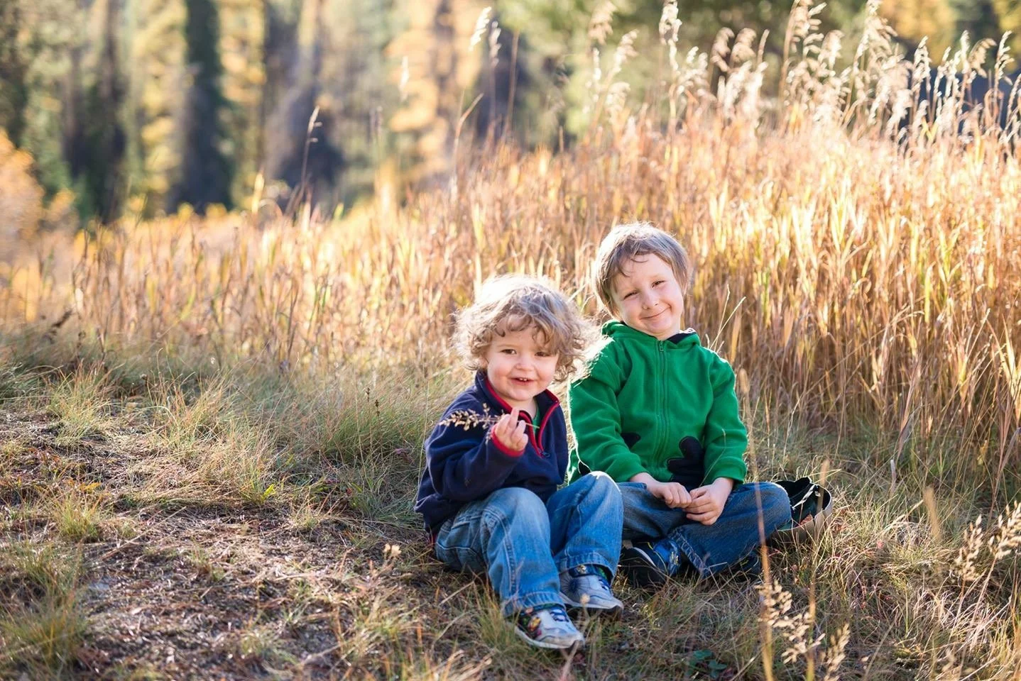 Two young boys sitting on the grass in a field of tall grass and autumn trees, smiling and enjoying the outdoor scenery.