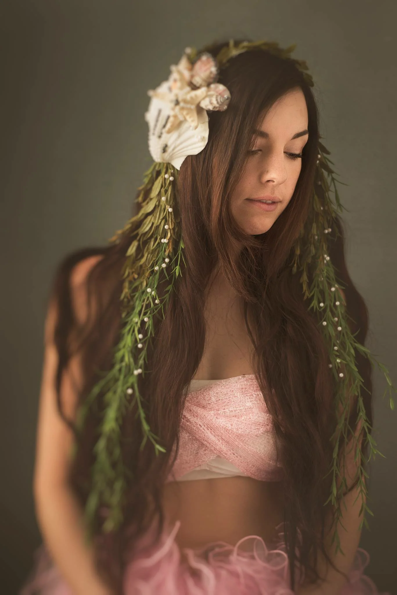 A young woman with long, wavy brown hair wearing a floral headpiece with seashells, shells, and greenery, and a pink strapless dress.
