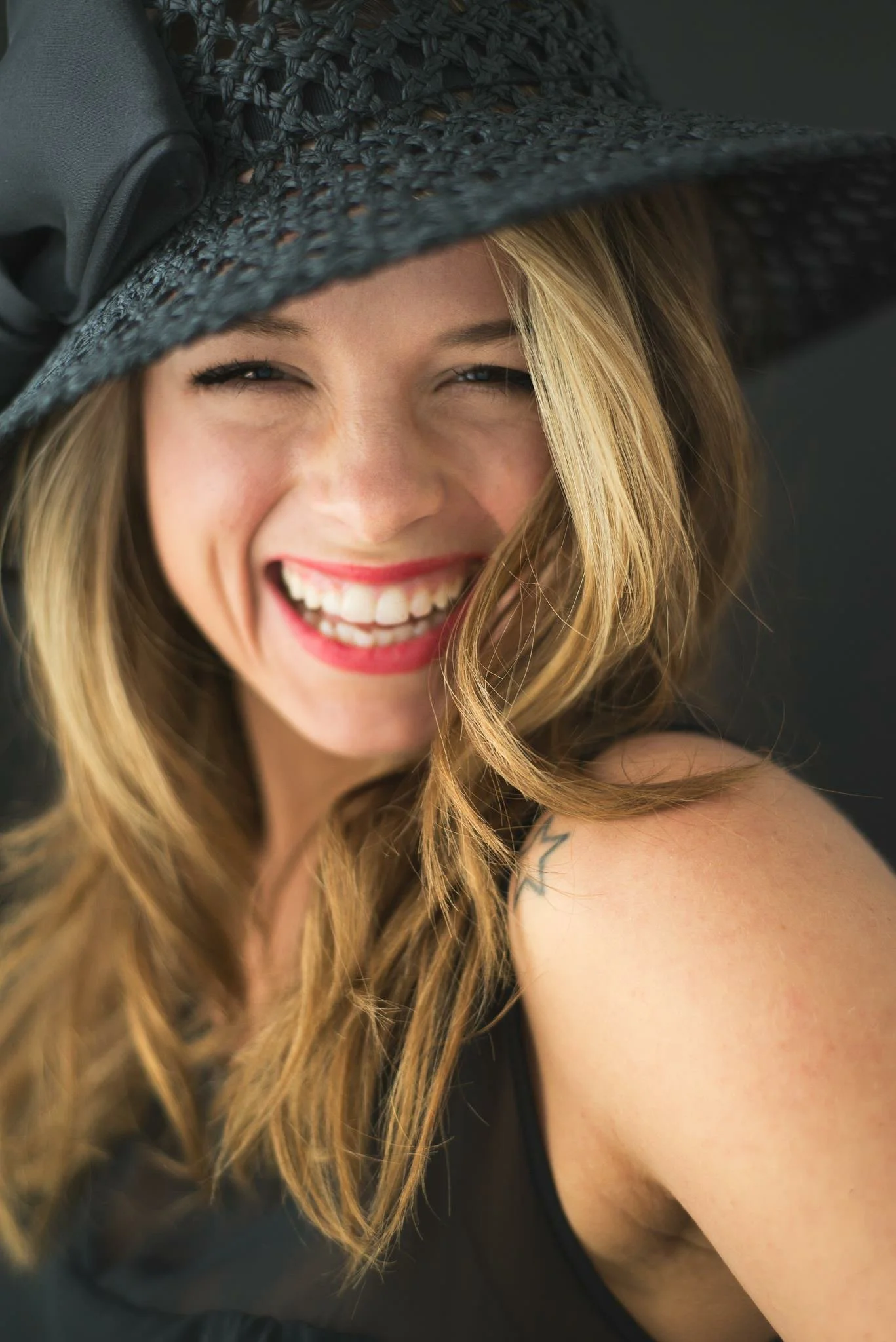 A woman smiling broadly, wearing a large black sunhat with a decorative bow, with long, wavy blonde hair, and a star tattoo on her shoulder.