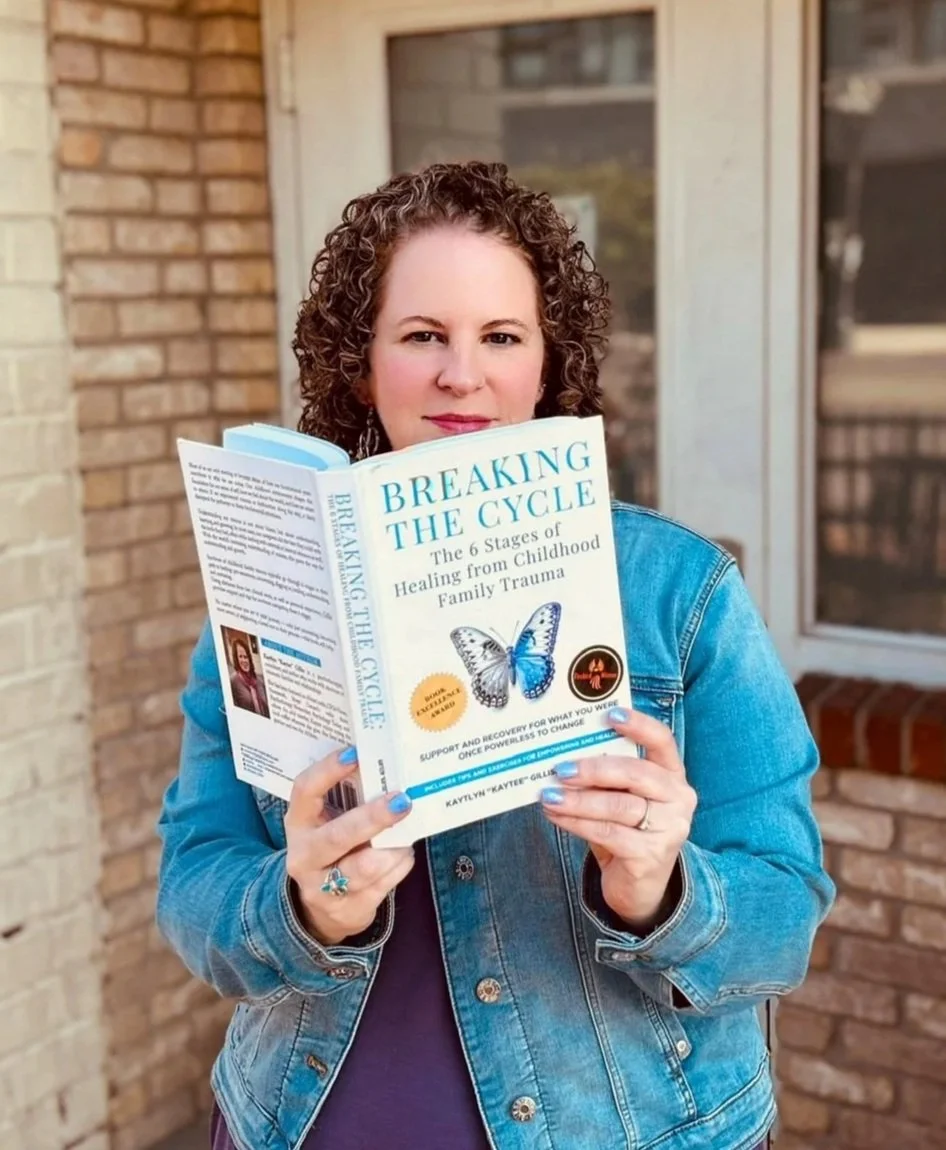A woman with curly hair wearing a denim jacket, holding a book titled 'Breaking the Cycle' in front of a brick house with large windows.