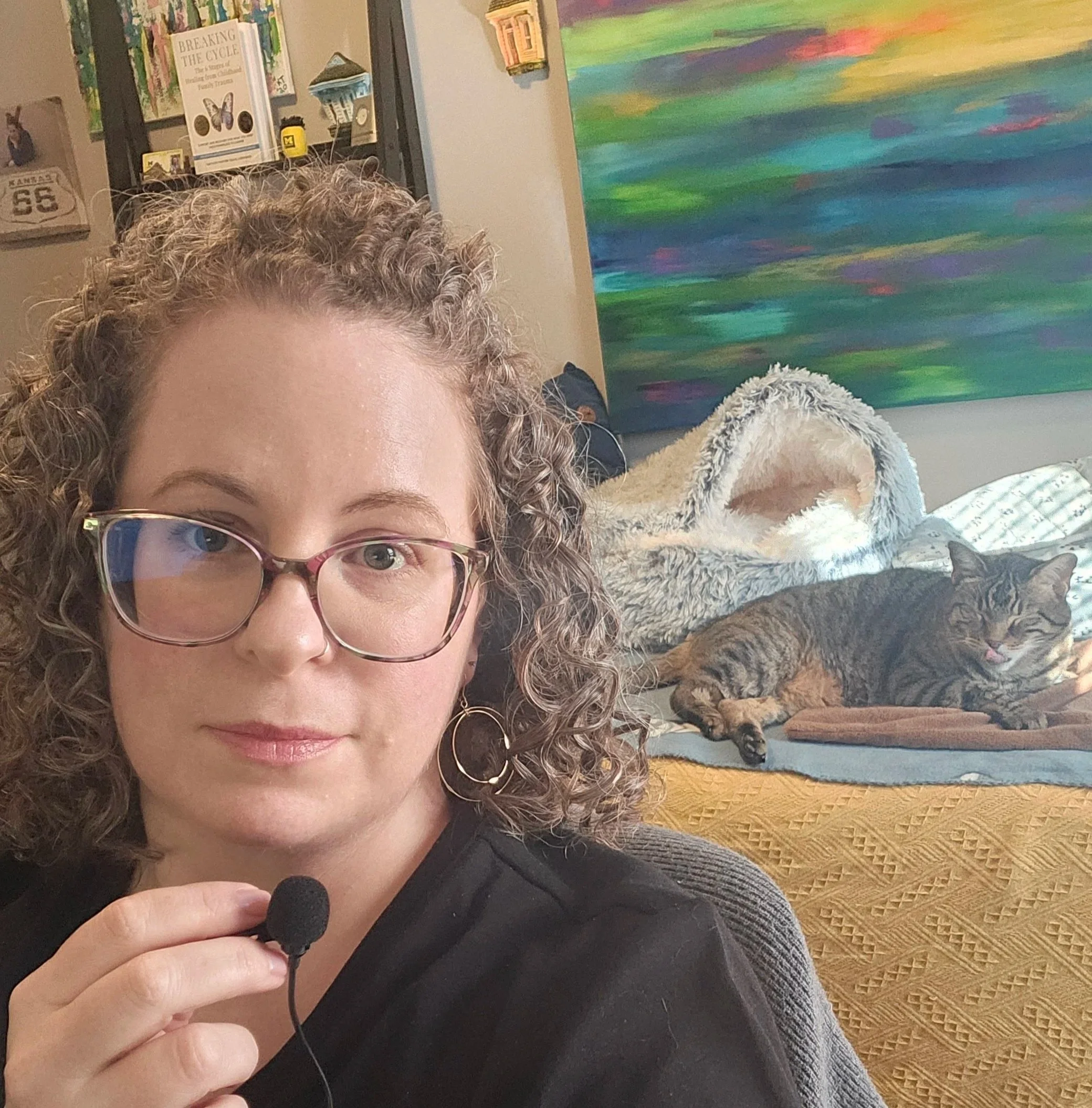 A woman wearing glasses and a black shirt with a gray knitted sweater, holding a microphone, with a painting of colorful abstract waves on the wall behind her.
