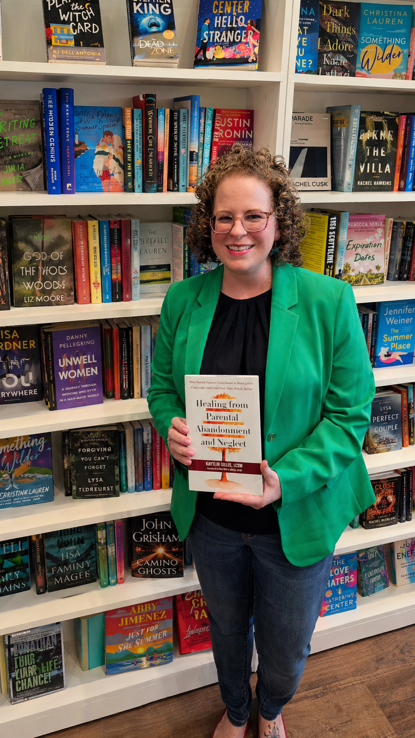 woman with curly hair holding a book in front of a bookcase in a bookstore. she is wearing a green blazer