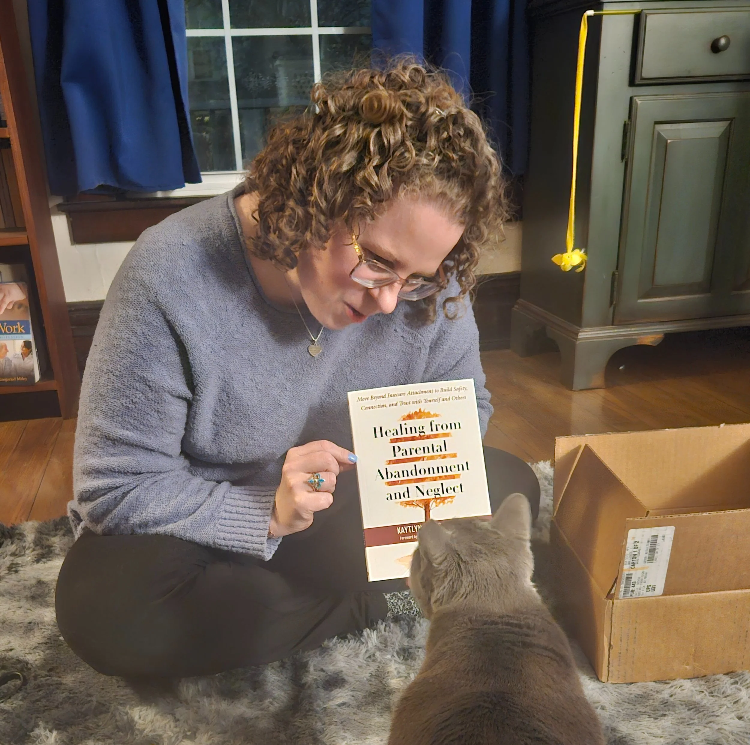 A woman with curly hair and glasses holding a book titled 'Healing from Parental Abandonment and Neglect' while a gray cat looks at her. They are sitting on a fluffy rug inside a room with wooden floors and a window behind.