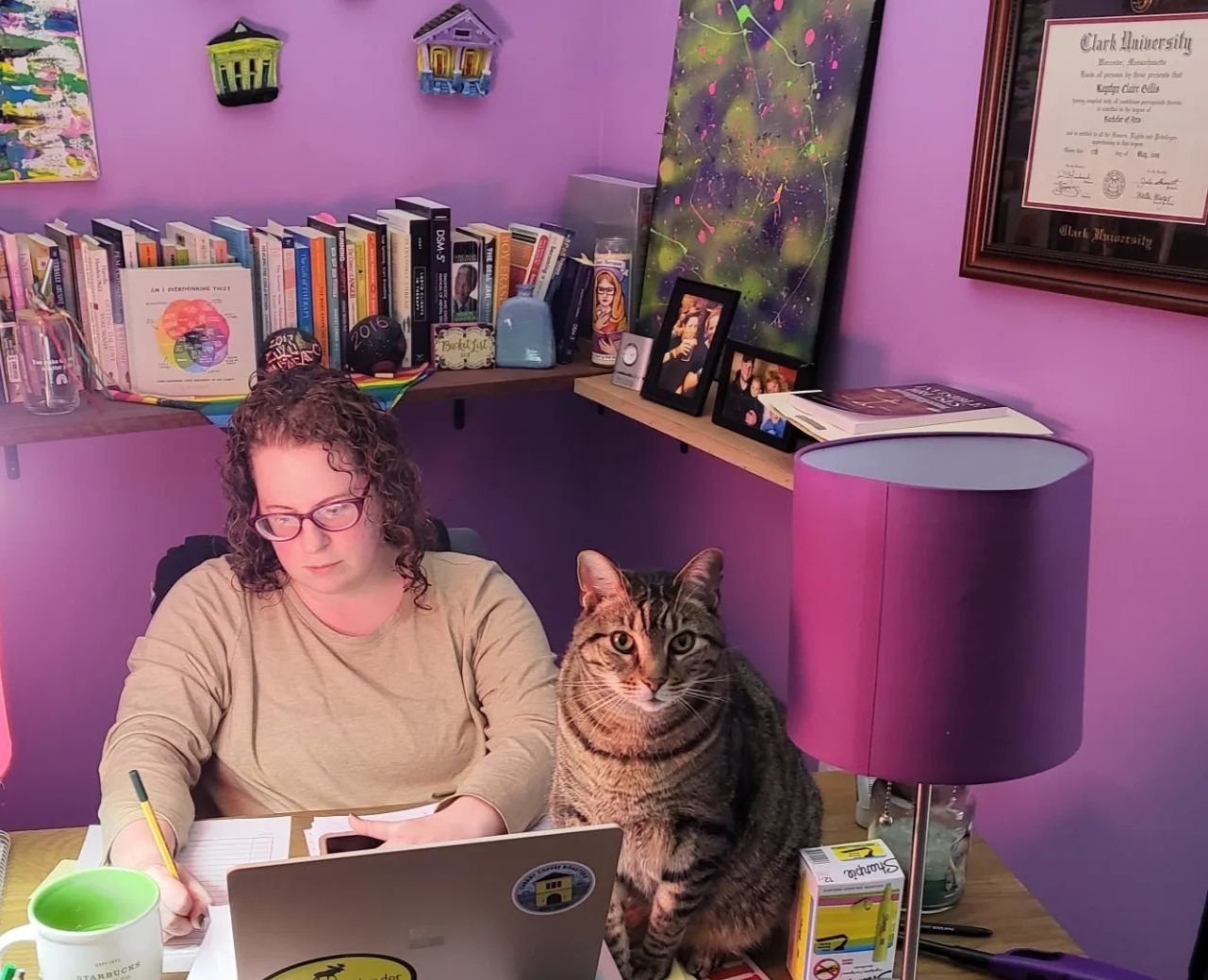 A woman with glasses working at her desk with a tabby cat sitting beside her, in a purple room with a bookshelf, framed photos, and art on the wall.
