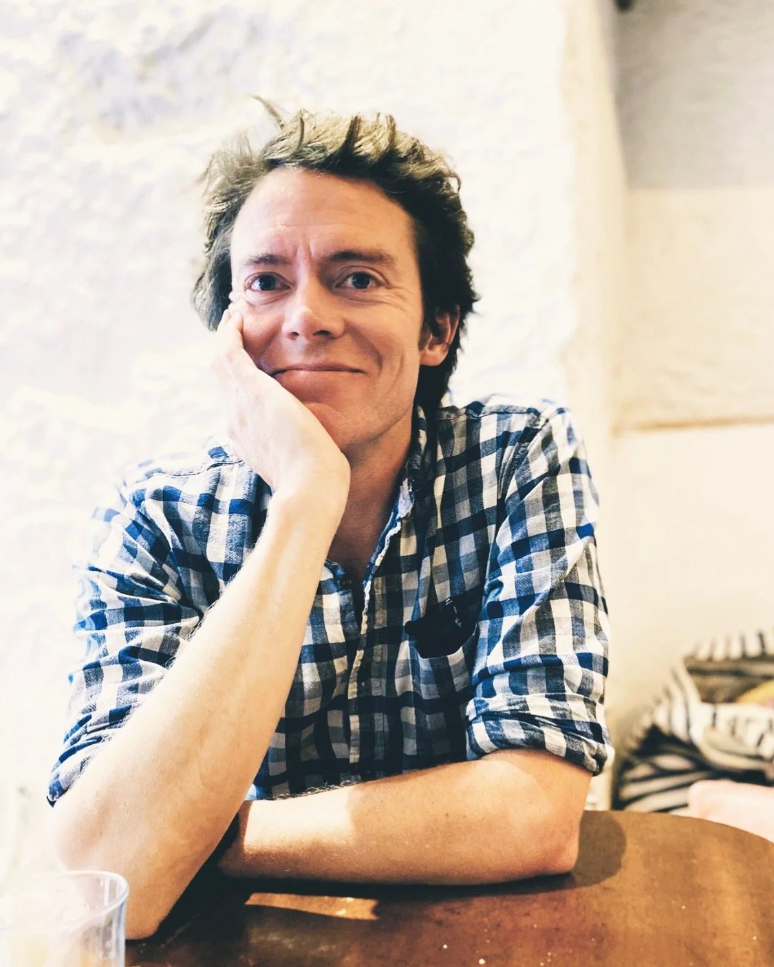 A man with dark, curly hair and light skin, resting his chin on his hand, wearing a blue and white checkered shirt, sitting at a table in a cozy indoor setting with a rough white brick wall in the background.