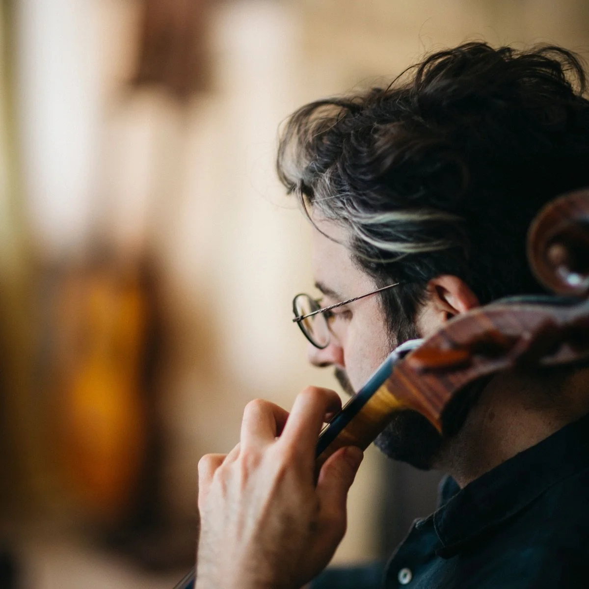 A man with dark hair, glasses, and a beard, resting his chin on a violin, appears to be deep in thought.