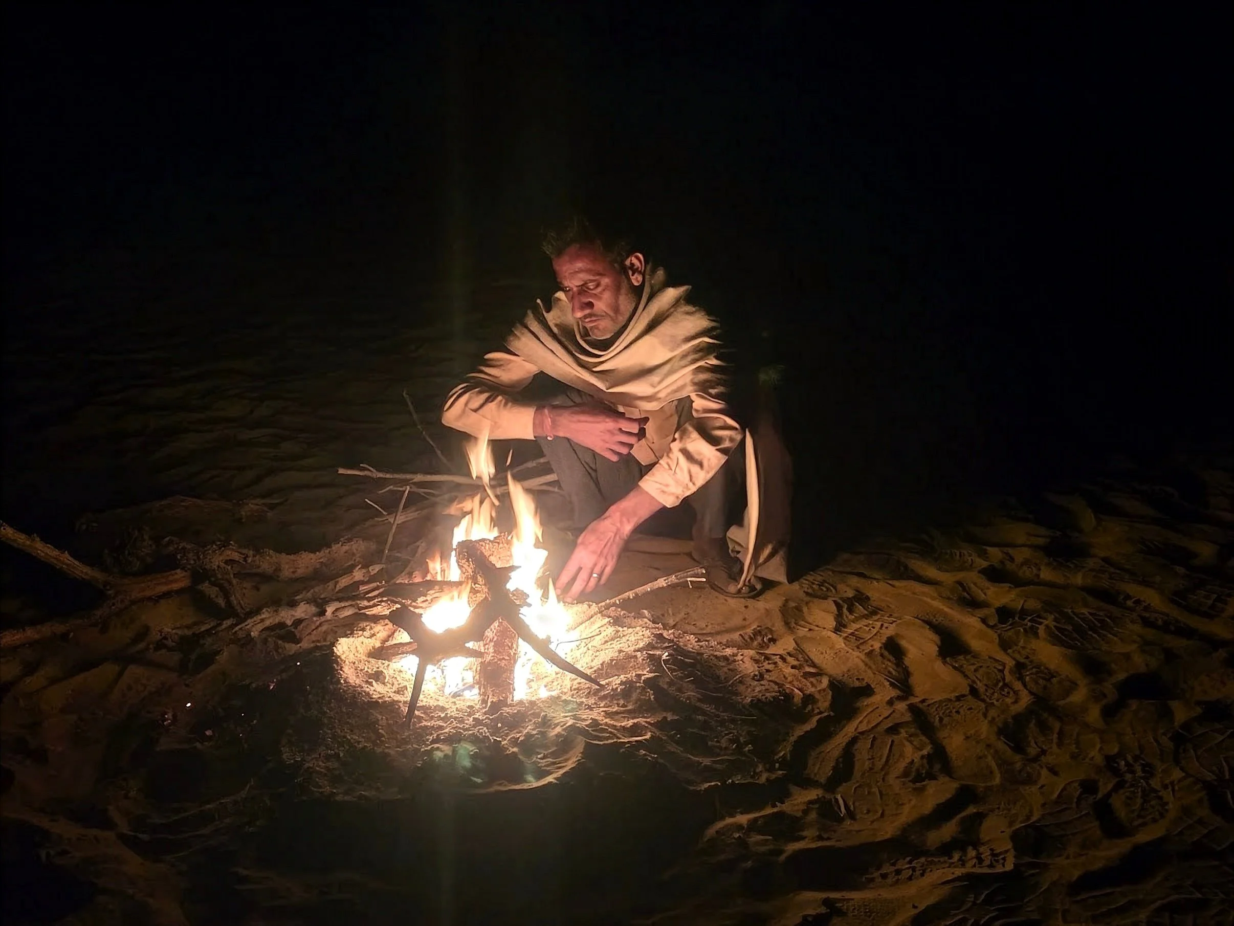 A man sitting on the beach at night, tending a small fire. The scene is dark with the focus on the fire and the man.