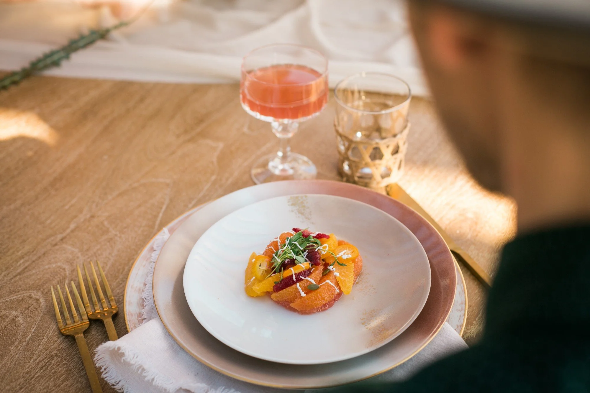 Plated citrus salad course with microgreens beside a cocktail at a sunlit table setting