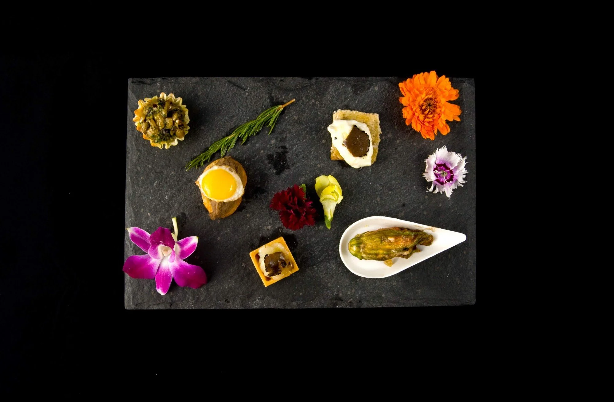 Overhead view of individually plated canapes and edible flowers arranged on black slate
